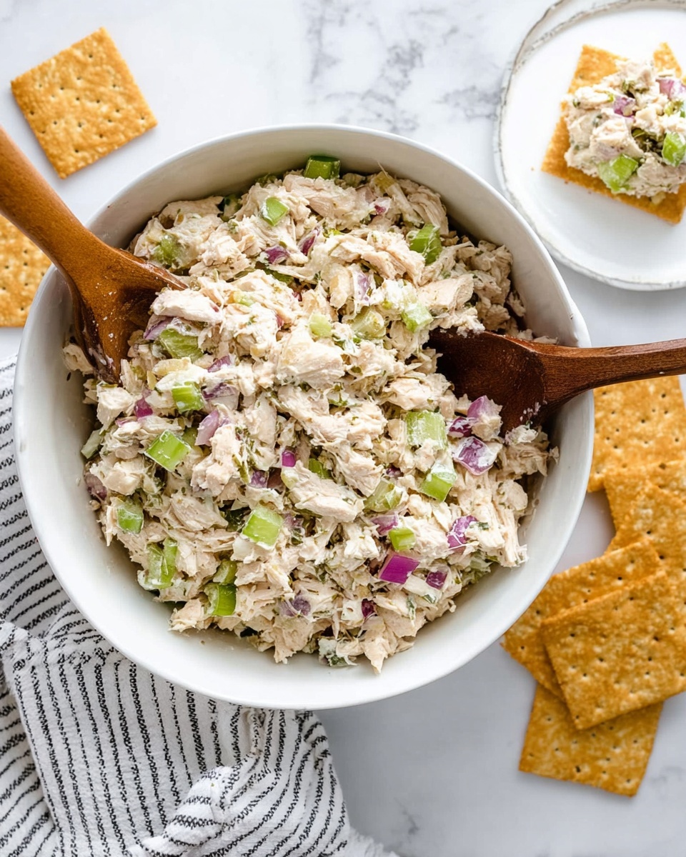 The image shows a close-up of a white bowl filled with a chunky light beige salad mixed with small pieces of green celery and bits of red onion throughout. A woman's hand holds a square golden-brown cracker topped with a scoop of this salad, displaying its creamy and textured mix. The background has a soft white marbled surface with more crackers blurred in the upper part of the image. The scene has bright natural lighting, highlighting the fresh and mixed ingredients photo taken with an iphone --ar 4:5 --v 7