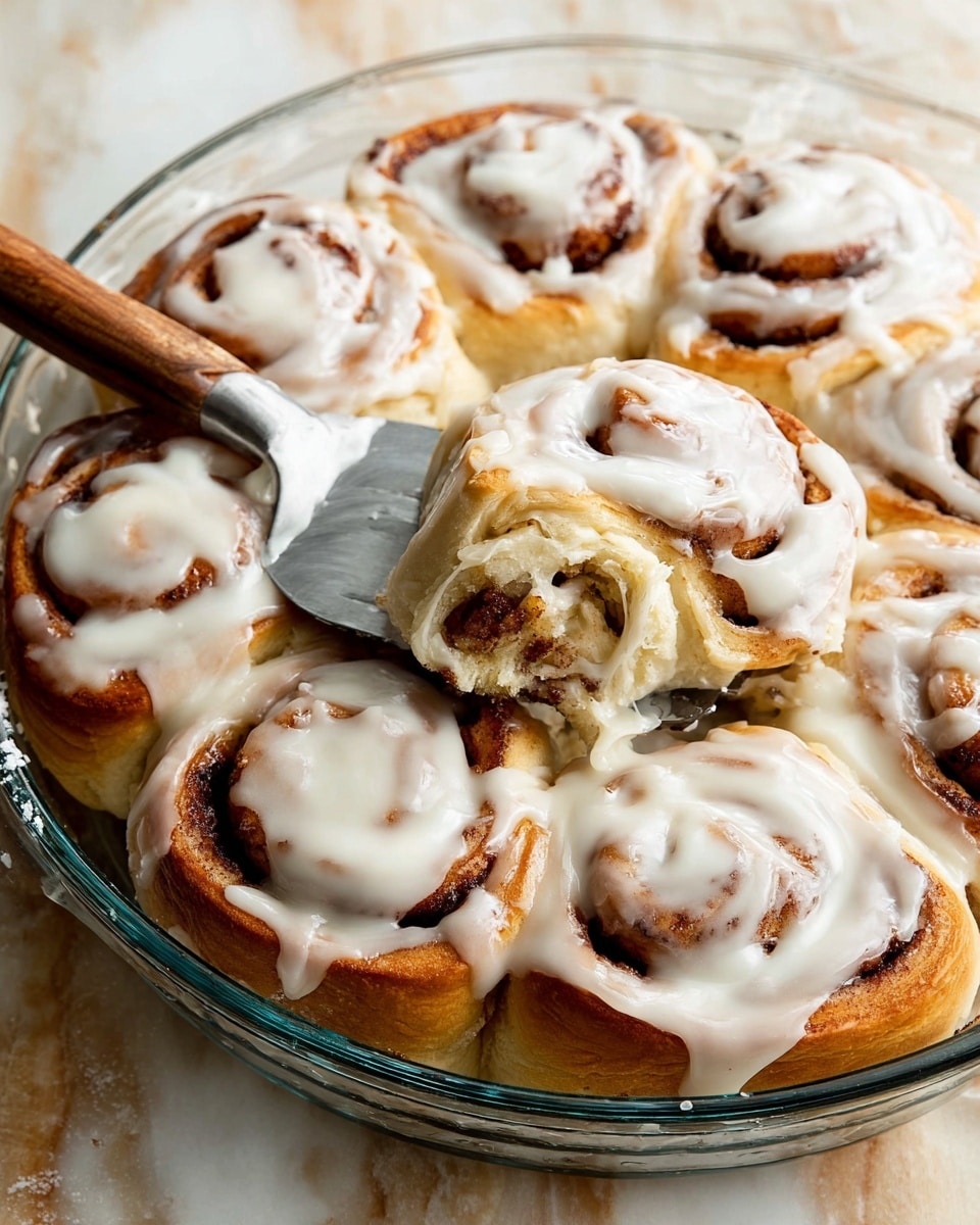 A clear round glass dish filled with eight cinnamon rolls, each thick and swirled with deep brown cinnamon layers and a soft golden-brown dough base, all topped with thick, creamy white icing that slightly drips over the edges. One cinnamon roll is being lifted with a metal spatula with a wooden handle, revealing its fluffy inside texture. The dish is placed on a white marbled surface that contrasts with the warm colors of the rolls. The whole scene looks fresh and inviting. photo taken with an iphone --ar 4:5 --v 7