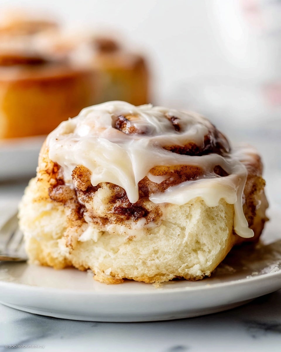 The image shows a close-up of a single cinnamon roll on a white plate, set on a white marbled surface. The cinnamon roll has three visible layers; the bottom layer is a soft, light beige dough base with a slightly golden crust. Above it is a middle layer thick with cinnamon swirls, dark brown in color with a textured, slightly sticky look. The top layer is creamy white icing spread thickly, dripping down the sides of the roll, smooth and glossy, covering the cinnamon swirls partially. The background is blurred, keeping the focus on the roll. Photo taken with an iphone --ar 4:5 --v 7