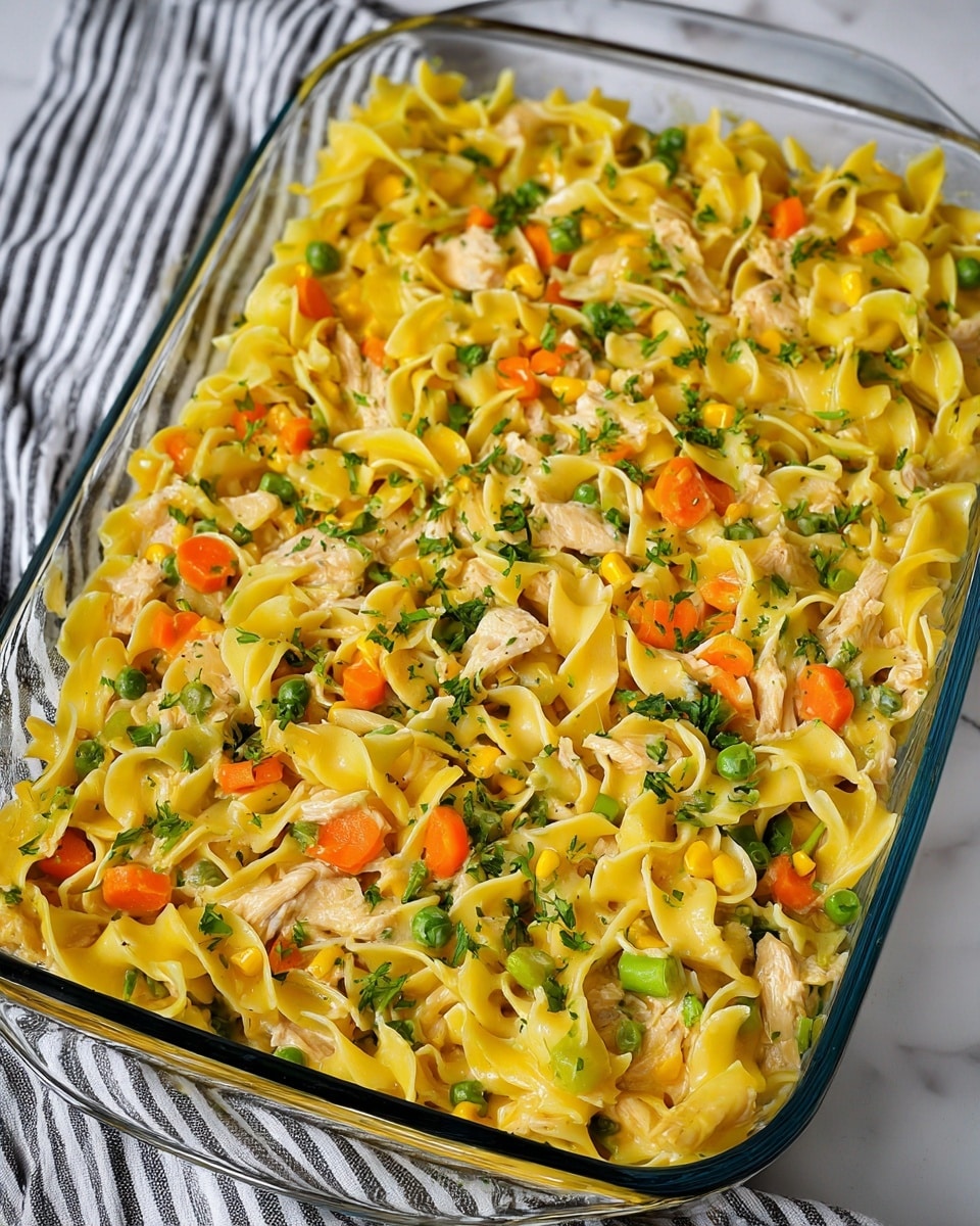 A close-up view of a glass baking dish filled with a creamy yellow egg noodle casserole mixed with green peas, orange carrot pieces, green beans, and chunks of cooked chicken. The noodles are soft and curled, coated evenly in a smooth sauce with small bits of herbs sprinkled on top. The dish sits on a white marbled surface with a striped cloth in the background. photo taken with an iphone --ar 4:5 --v 7