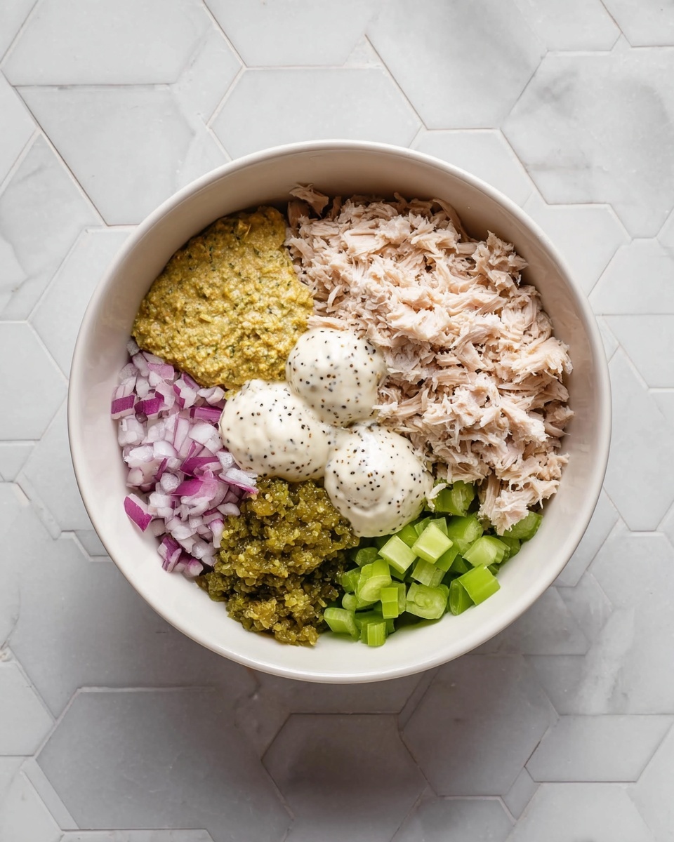 A white bowl sits on a white marbled hexagon tile surface, filled with several layers of ingredients separated neatly. On the right side, there is a large layer of light beige shredded tuna. To the bottom right, small pale green diced celery pieces form a cluster, with finely chopped red onion in the bottom left corner. Next to the onion and slightly above, there is a small pile of green relish. Centered on top of these layers are three dollops of creamy sauces: a white mayonnaise-like sauce with black pepper, a darker yellow mustard sauce, and another white creamy sauce, all placed side by side. The photo taken with an iphone --ar 4:5 --v 7