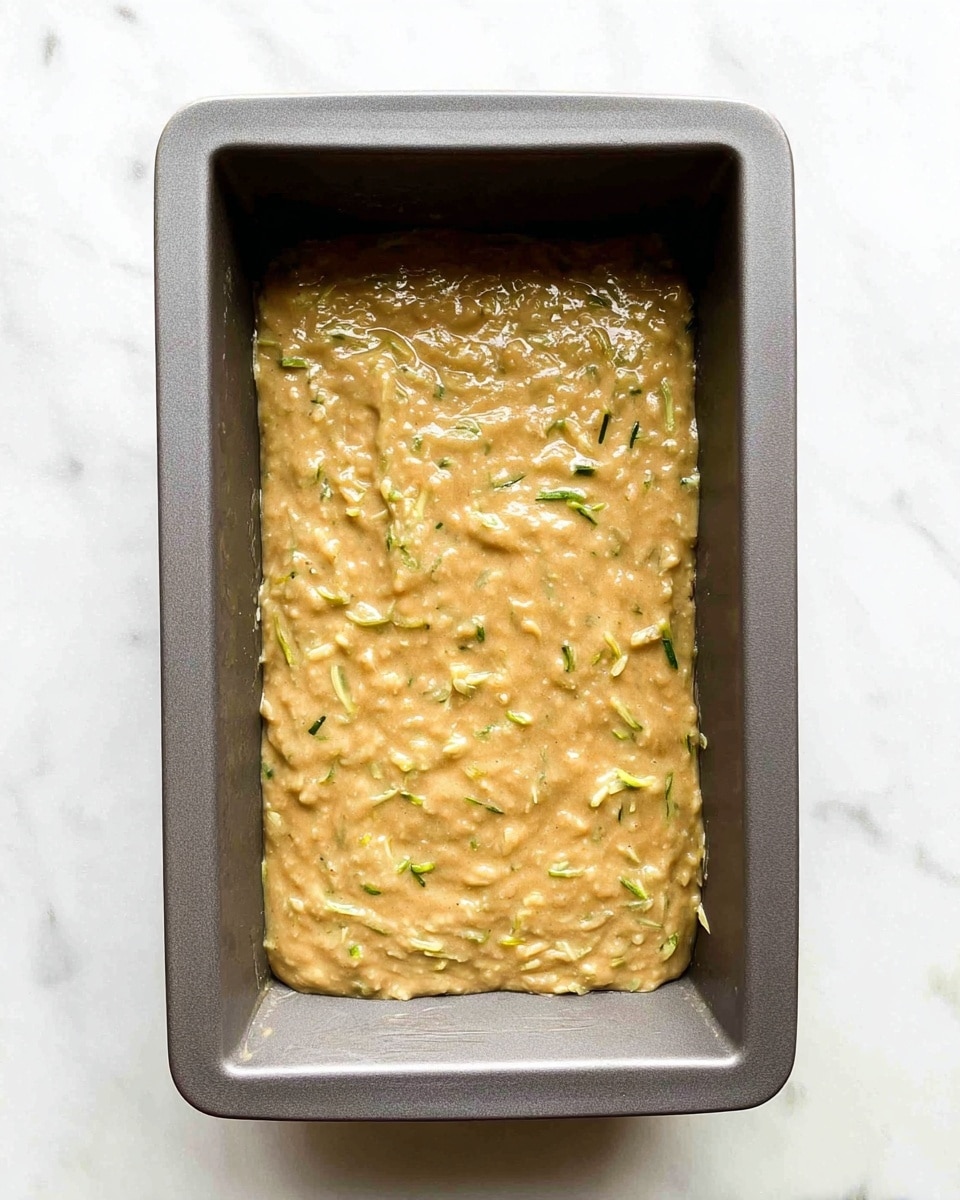 A gray rectangular baking pan is filled with thick, light brown batter that has visible small pieces of shredded green vegetables spread evenly throughout. The batter’s texture looks smooth with some soft lumps, and it covers the bottom of the pan in one even layer. The pan is placed on a white marbled surface, which provides a clean and bright background. Photo taken with an iphone --ar 4:5 --v 7