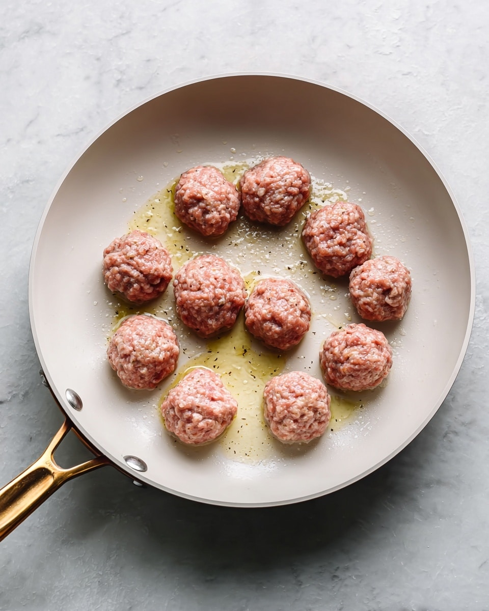 A white frying pan with a gold-colored handle holds twelve uncooked meatballs. The meatballs are arranged mostly in a loose circle with some inside the circle, evenly spaced out. They have a pink, raw look with a rough texture showing bits of white inside. Around the meatballs, there is light golden oil sizzling in the pan. The pan sits on a white marbled surface. photo taken with an iphone --ar 4:5 --v 7