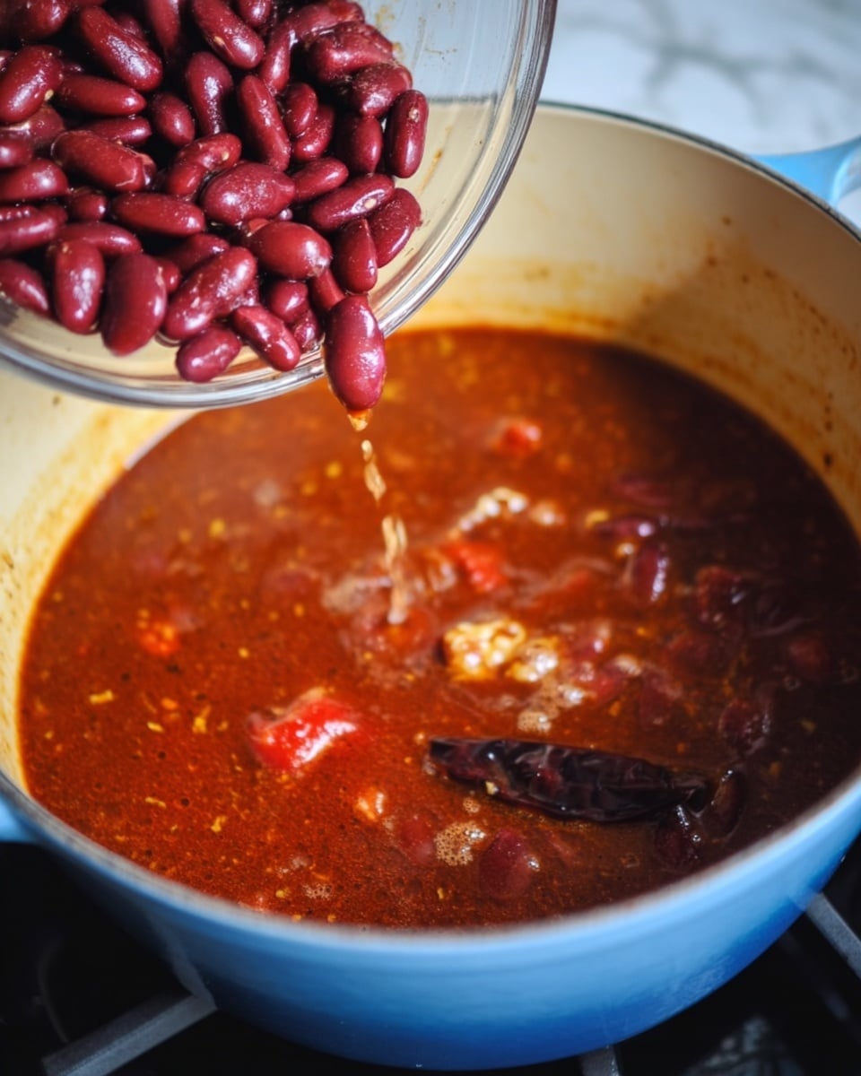 A close-up image shows a pot filled with a rich, bubbling red-brown stew with visible tomato pieces and a dried chili floating on the surface. A woman's hand is pouring shiny dark red kidney beans from a transparent bowl into the stew. The pot is cream-colored inside with a blue outer rim, sitting on a black stove, while the background is a white marbled texture. The colors mix warm reds and browns with the glossy kidney beans about to sink into the stew, creating a hearty and thick look. Photo taken with an iphone --ar 4:5 --v 7