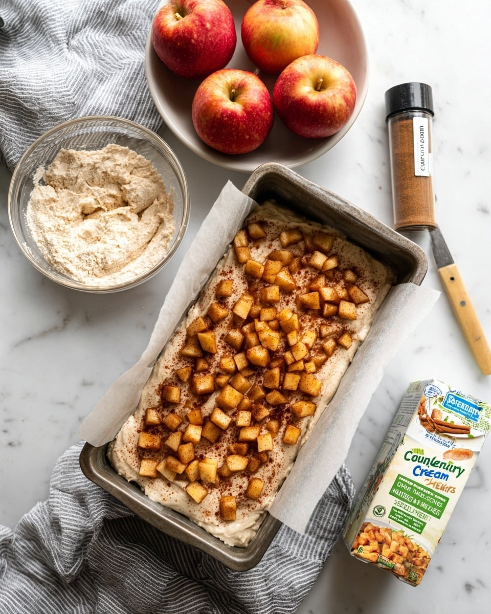 A clear glass mixing bowl sits centrally on a white marbled surface, filled with a creamy, light beige batter with a thick and slightly rough texture, swirled in the middle with a white spatula resting inside it. To the left, a white bowl is filled with several red apples with yellow and green hues, partially wrapped by a blue and white striped cloth. On the lower left, a stick of butter in blue and yellow wrapping is partly visible. To the right of the bowl, a small glass bowl contains chopped apple pieces coated with cinnamon, showing light brown and beige tones. Nearby, a small bottle of ground cinnamon with a green label stands next to a knife with a wooden handle. At the top right corner, a carton of cage-free large eggs with blue and white packaging is positioned. Photo taken with an iphone --ar 4:5 --v 7