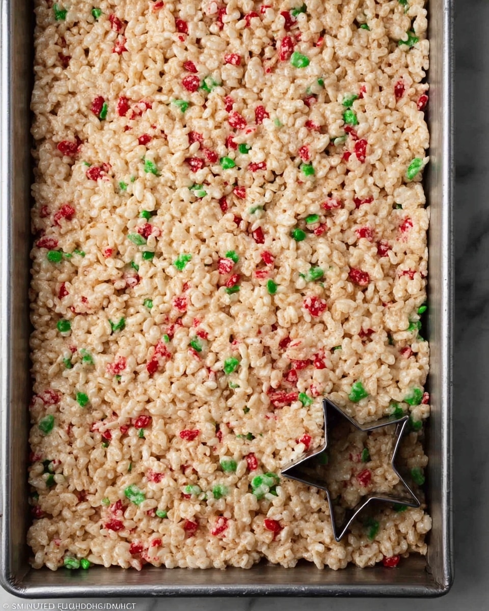 A rectangular metal tray is filled with a thick, even layer of light tan crispy rice treats mixed with scattered small red and green candy pieces throughout. On the lower right side of the tray, a metal star-shaped cookie cutter is pressed into the treats, slightly cutting into the top layer. The texture looks crunchy and sticky with the colorful candies adding small pops of color on the soft, clustered rice cereal base. The background features a white marbled surface photo taken with an iphone --ar 4:5 --v 7