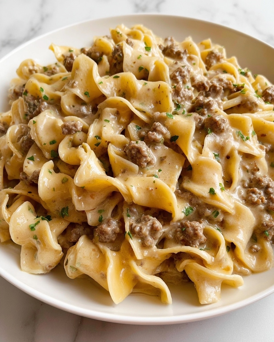 The image shows a close-up of a white plate filled with a creamy pasta dish featuring several layers of wide, twisted egg noodles coated in a light beige sauce. Mixed through the pasta are small chunks and crumbles of browned ground meat, giving a slightly rough texture mixed with the smooth and glossy look of the sauce. The sauce has a rich, creamy appearance with tiny flecks of green herbs scattered throughout, adding subtle color contrast. The noodles have soft folds and wavy edges that catch the light, showing a shiny, tender surface. The background is a white marbled texture. photo taken with an iphone --ar 4:5 --v 7