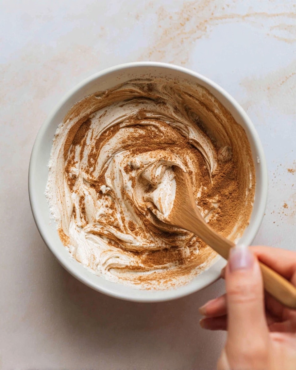 A close-up view of a white bowl held by a woman's hand shows a mix of creamy white and light brown swirled together inside. The creamy part has a thick, smooth texture, while the brown appears as fine powder sprinkled and partly mixed around the edges and center. A wooden spoon is stirring the mixture, creating soft, wavy patterns. The background is a white marbled texture. photo taken with an iphone --ar 4:5 --v 7