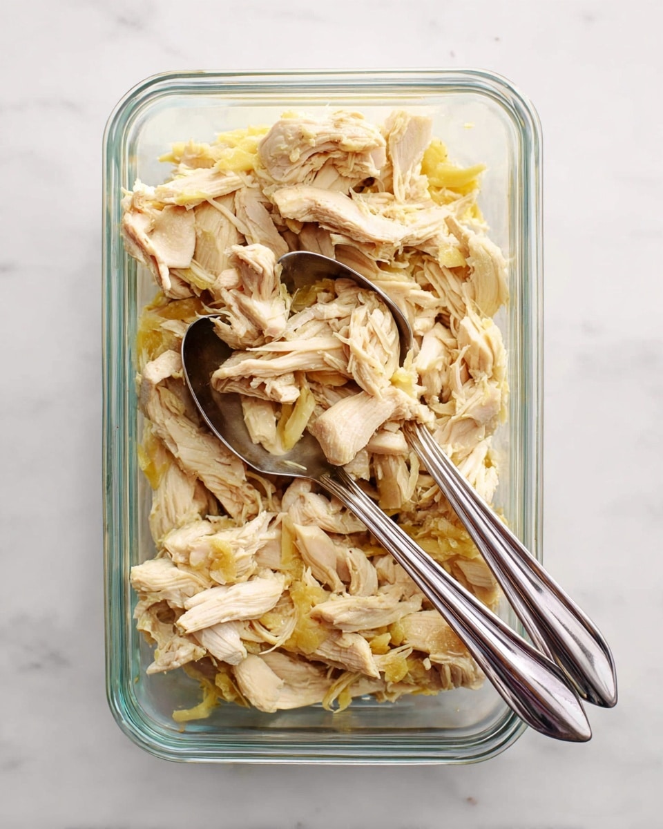 A clear glass rectangular container filled with shredded pale beige and yellow cooked chicken pieces, showing soft, fibrous texture with some skin still attached in yellow color; two silver serving spoons rest inside at the bottom of the container, angled outward; the container is placed on a white marbled surface. photo taken with an iphone --ar 4:5 --v 7