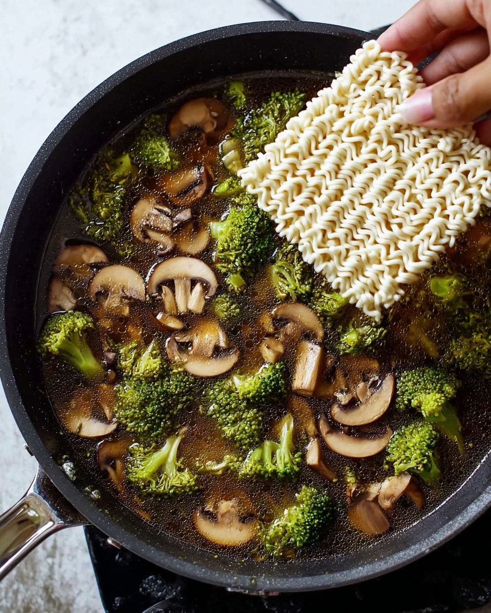 A close-up view of a black frying pan filled with dark brown broth, floating with green broccoli florets and sliced brown mushrooms scattered throughout the liquid. A woman's hand is seen lowering a block of white instant ramen noodles with a wavy texture into the broth from the top left corner. The pan sits on a white marbled surface with part of the metallic pan handle visible on the left side. Photo taken with an iphone --ar 4:5 --v 7