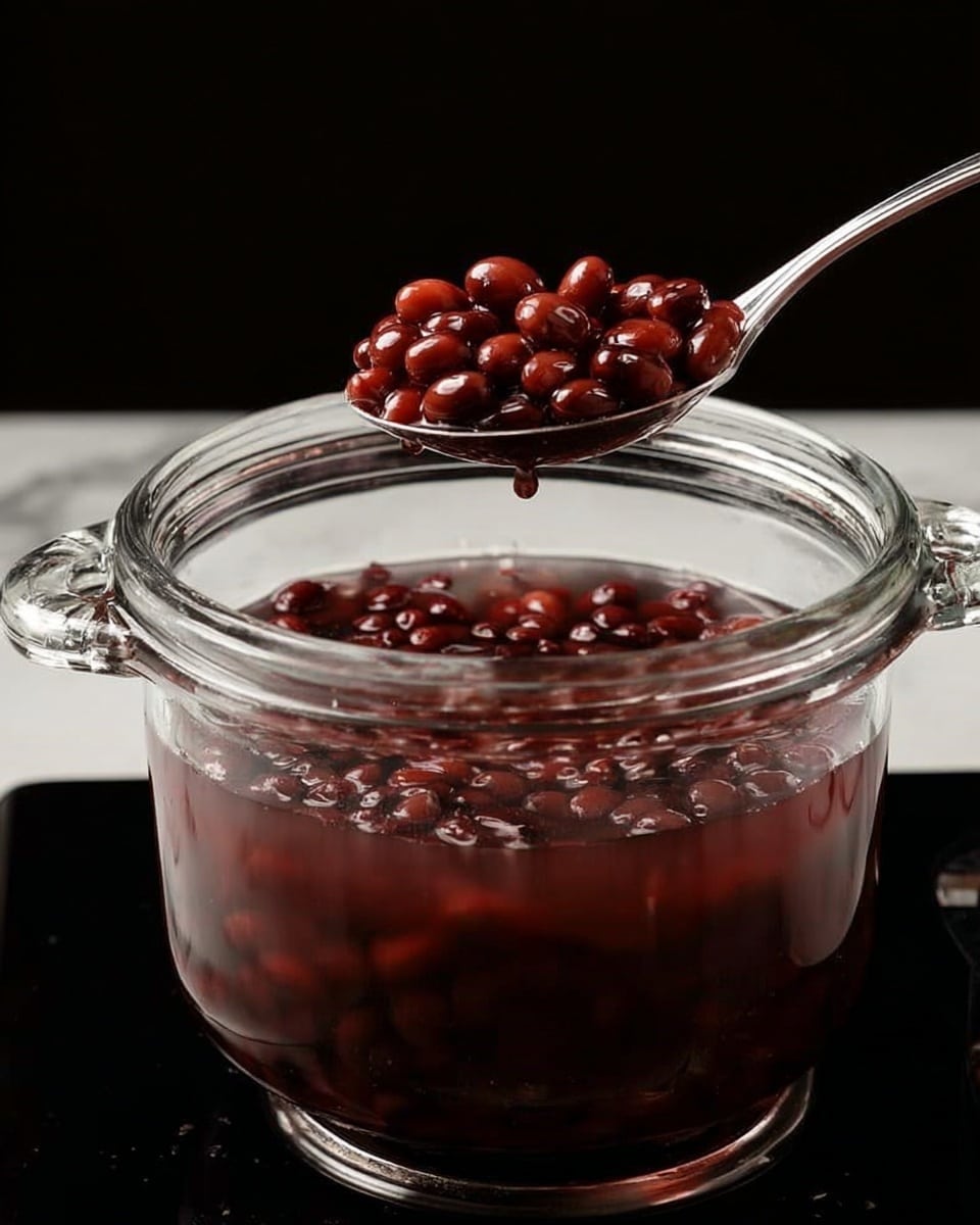 The image shows a clear glass pot filled with dark red beans soaking in water, with a silver spoon lifting a portion of the beans above the pot. The pot has two small handles on each side and is placed on a black stove. The background is black, and the surface underneath the stove is changed to a white marbled texture. The beans look smooth and shiny, and the water inside the pot is slightly tinted by the beans. Photo taken with an iphone --ar 4:5 --v 7