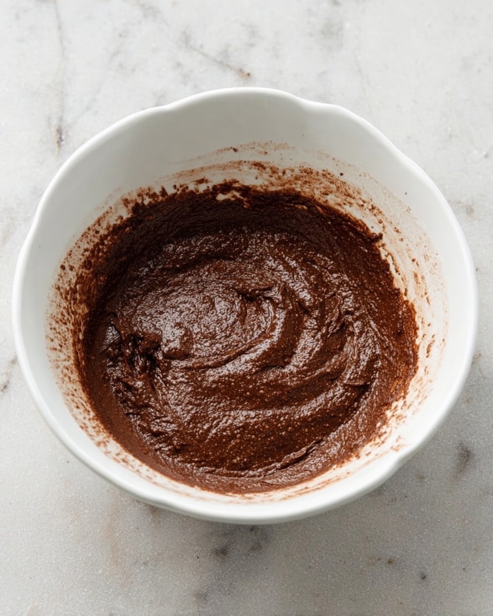 A white bowl with a slightly scalloped edge holds thick chocolate batter that is dark brown and slightly shiny with a rough texture. The batter is spread unevenly inside the bowl, with visible mixing marks and a bit of batter stuck to the sides near the rim. The bowl is placed on a white marbled surface. photo taken with an iphone --ar 4:5 --v 7