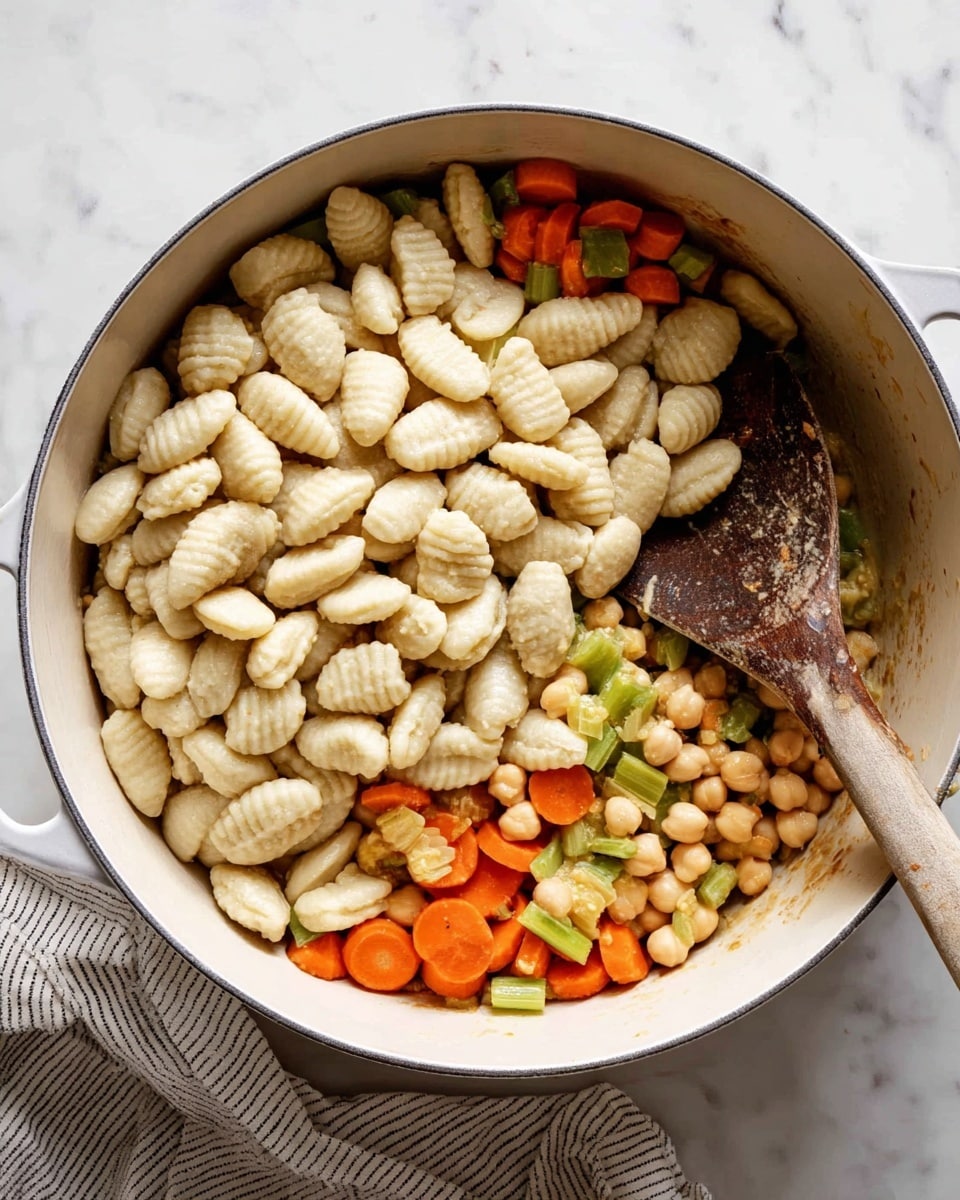 In a white round pot, there are three main layers visible: at the bottom, a mix of cooked vegetables including small orange carrot slices, pale green celery pieces, and chickpeas that are light beige and round, sitting loosely; on top of these, a large pile of raw gnocchi, pale beige with soft ridges visible on each piece, unevenly spread covering about half of the vegetables; a wooden spoon with a dark brown, weathered look rests partly in the pot, its handle pointing outwards to the top right. The pot sits on a white marbled surface with a striped cloth nearby. Photo taken with an iphone --ar 4:5 --v 7