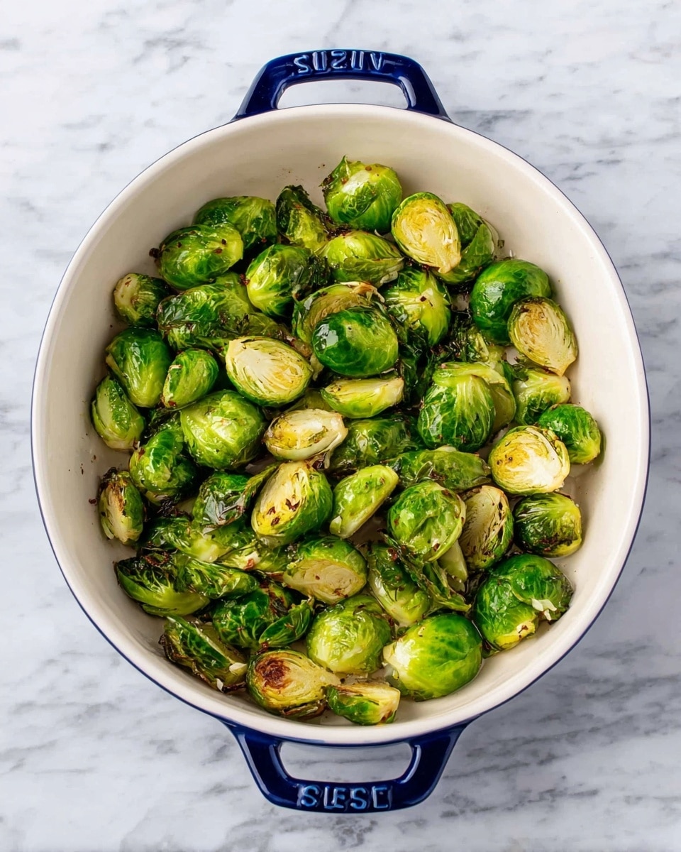 The image shows a white oval baking dish with dark blue handles on a white marbled surface, filled with roasted Brussels sprouts. The Brussels sprouts are cut in half, showing a mix of bright green leafy textures and slightly browned edges from cooking. They are arranged evenly across the dish, with a few browned spots and dimples adding a roasted effect, and light oil or seasoning visible on the surface. photo taken with an iphone --ar 4:5 --v 7