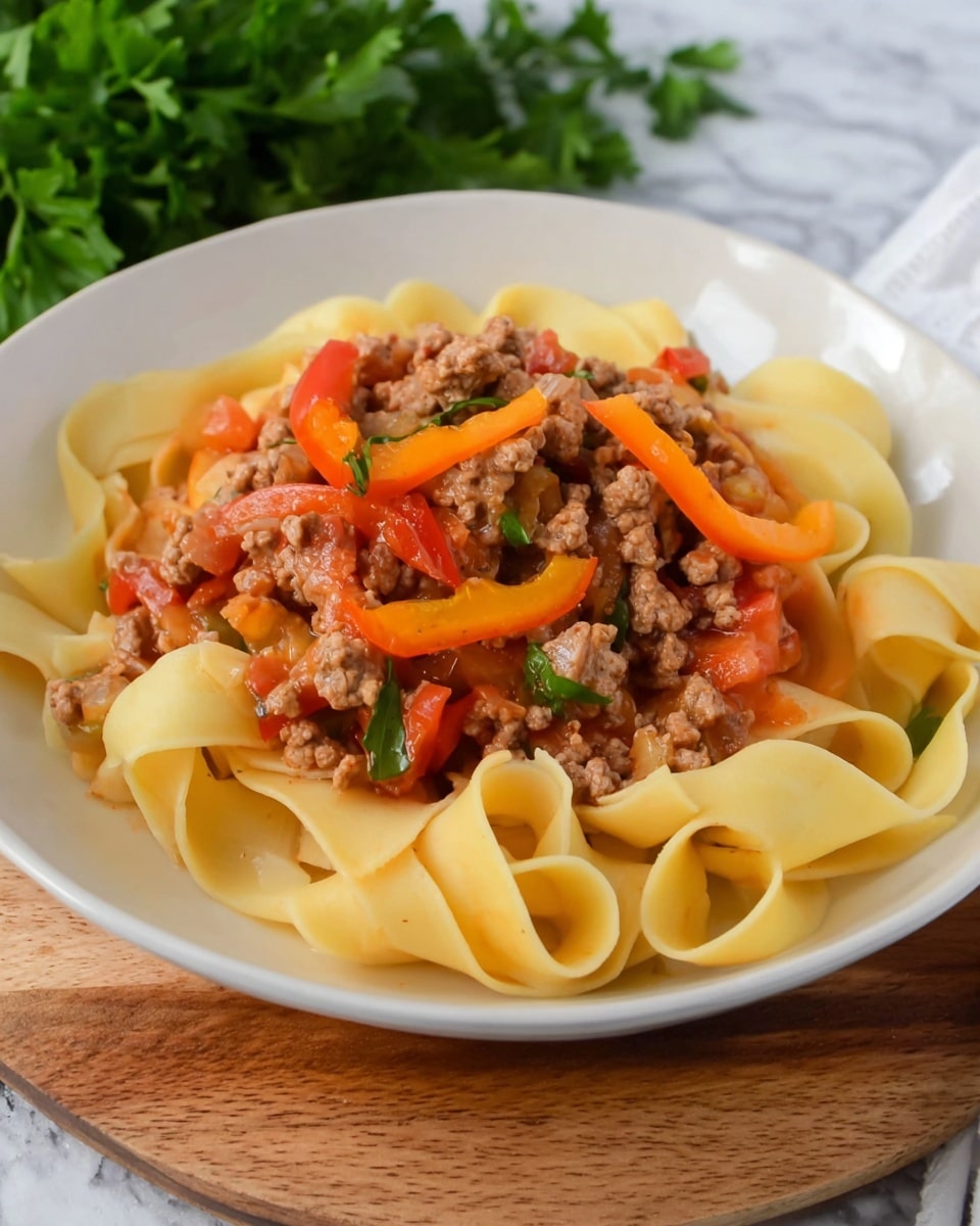 A close-up view of wide, pale yellow flat pasta noodles layered evenly in a black pan with a mix of light brown cooked ground meat scattered throughout. Bright orange and red thin bell pepper strips add color on top of and around the pasta. Small pieces of red diced tomatoes and bits of green herbs are spread evenly over the dish, adding more texture and detail. The pan rests on a white marbled surface with a beige spoon and some green herbs visible near the edge. photo taken with an iphone --ar 4:5 --v 7