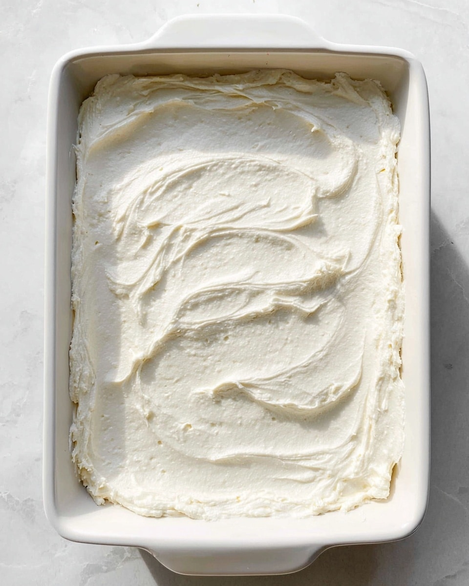The image shows a white square baking dish filled with a thick, evenly spread layer of creamy white mixture. The creamy layer has a smooth texture with some gentle swirls and small uneven patches, giving it a soft, slightly whipped appearance. The edges of the creamy layer are slightly rough where it meets the dish’s sides. The dish is placed on a white marbled surface, and the lighting highlights the creamy texture's subtle shadows and light reflections. photo taken with an iphone --ar 4:5 --v 7