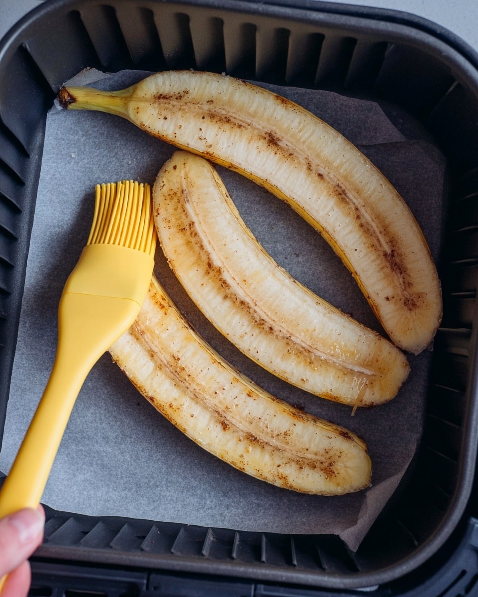 The image shows three long yellowish banana halves with brown spots lying inside a black air fryer basket lined with gray parchment paper. A yellow silicone brush held by a woman's hand is spreading some liquid over the top of one banana half. The bananas are peeled and placed with the cut side facing up, showing their soft, pale yellow flesh with a slight brown speckled texture. The black sides of the air fryer basket frame the bananas closely. photo taken with an iphone --ar 4:5 --v 7