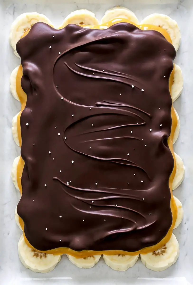A close-up view of a layered dessert on a white marbled surface, showing three main layers: the bottom layer is made of round, thick yellow banana slices, arranged side by side forming a base; the middle layer is a smooth, shiny caramel with a light golden-brown color that covers the bananas evenly; the top layer is a thick, glossy dark chocolate spread with visible waves and folds, sprinkled lightly with coarse white sea salt. photo taken with an iphone --ar 4:5 --v 7