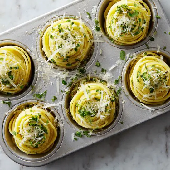 A metal muffin tray with six circular molds, each filled with thin, round slices of yellow dough stacked in layers, about 4-5 layers per mold. The dough layers show a smooth texture with small green herb pieces sprinkled on top and around the edges. Light bits of grated white cheese or garlic are scattered over the dough and inside the molds. The background is a white marbled surface. Photo taken with an iphone --ar 4:5 --v 7
