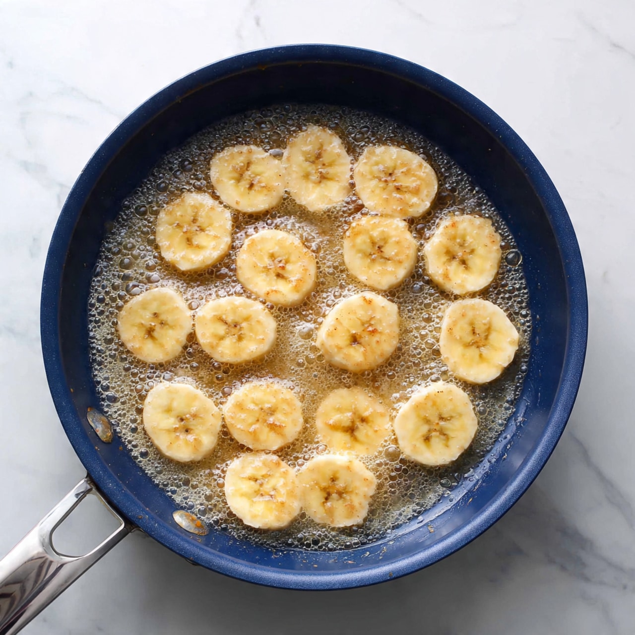 A dark blue frying pan filled with one layer of light yellow banana slices, each slice thin and round, cooking in a bubbling light brown syrup spread evenly beneath them. The frying pan sits on a white marbled surface, showing some bubbles rising in the syrup and slight caramelization on a few slices, with the pan handle extending to the left side. The scene is well-lit, with soft natural light highlighting the shiny syrup and smooth texture of the banana slices. Photo taken with an iphone --ar 4:5 --v 7