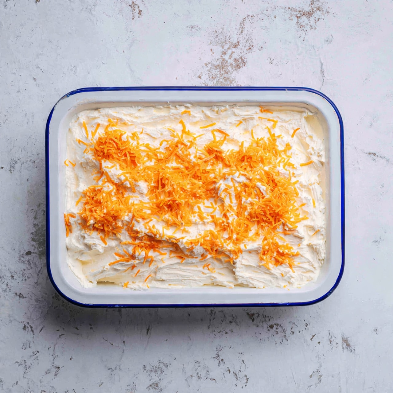 A white rectangular enamel dish with a thin blue rim is filled with a thick, creamy white layer that looks smooth but with some soft lumps. On top, there is a generous sprinkling of shredded orange cheese spread unevenly across the surface with some small clusters. The dish sits on a white marbled textured surface. photo taken with an iphone --ar 4:5 --v 7