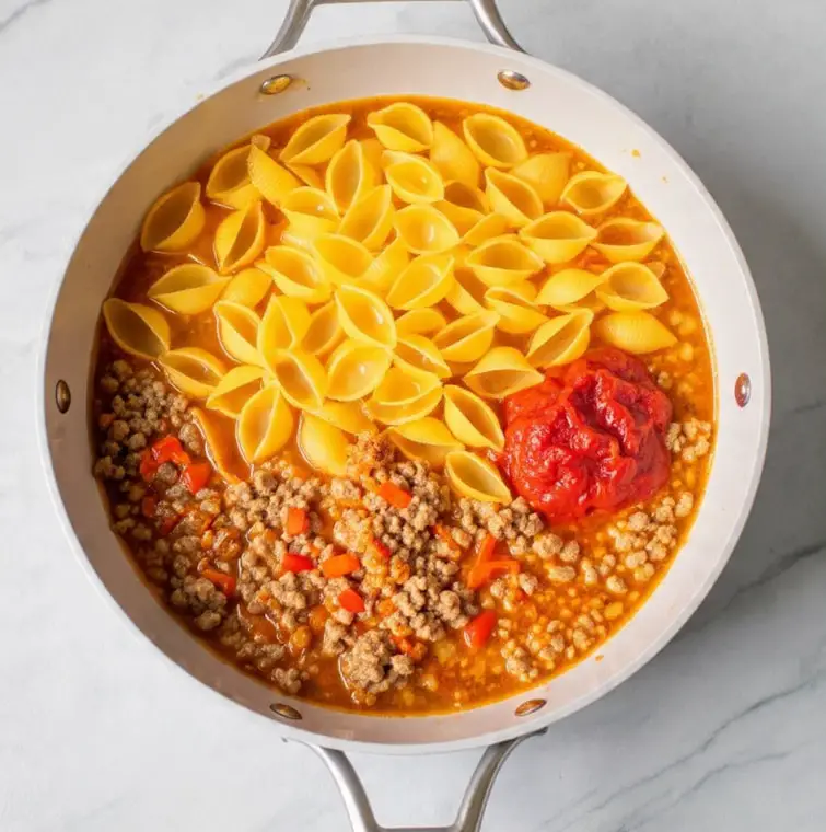 A white pan with two silver handles holds three main layers of food. The bottom layer is a golden-brown ground meat mixed with small pieces of red bell pepper and some sauce. Above that is a thin layer of orange-red broth that fills most of the pan. On top of everything is a layer of uncooked large yellow shell pasta, arranged loosely and spread across the center and right side of the pan. Near the pasta, there is one small clump of red tomato paste resting on the broth. The pan sits on a white marbled surface. photo taken with an iphone --ar 4:5 --v 7