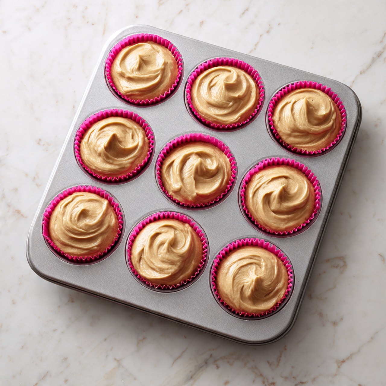 A metal muffin tray with 12 round cavities filled with light brown cupcake batter sitting in pink paper liners, the batter is smooth and slightly mounded in each cavity with soft peaks and swirls on top. The tray is placed on a white marbled surface, showing clear grooves and a shiny texture. Photo taken with an iphone --ar 4:5 --v 7