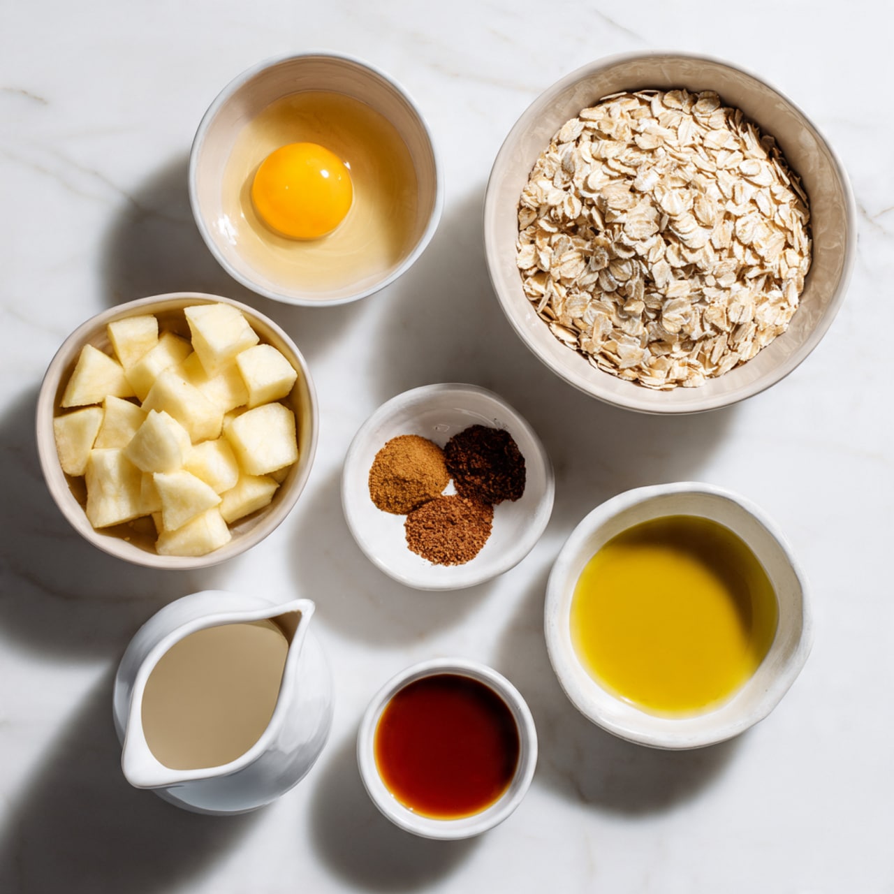 Seven white bowls and a white jug are arranged on a white marbled surface, each holding a different ingredient. The largest bowl at the top right is full of old fashioned oats, off-white with a slightly rough texture. To its left, a smaller bowl holds a raw egg with a bright yellow yolk floating in clear egg white. Below the oats is a bowl of peeled, cubed apples showing cream and light red tones. Near the center is a tiny bowl with three brown spice powders—cinnamon, nutmeg, and ground ginger—grouped but separate. Underneath, a small bowl contains a shiny pool of reddish-brown maple syrup, while to the bottom left a white jug holds smooth, creamy almond milk. Lastly, a small labeled bowl near the bottom right is filled with golden olive oil that shines in the light. The setup is simple and neat. photo taken with an iphone --ar 4:5 --v 7