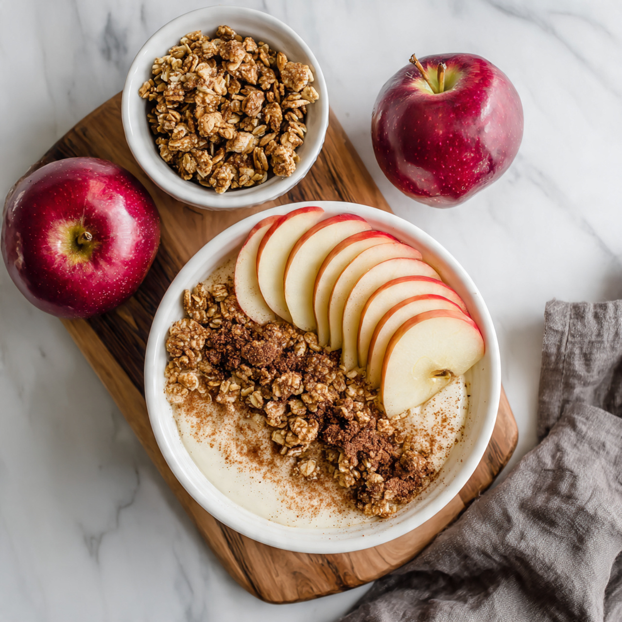 A white bowl sits on a wooden board on a white marbled surface, filled with a creamy white base as the bottom layer. On top of this, there is a layer of chunky granola scattered unevenly, and thin, evenly spaced slices of red apple with a pale yellow inside slide across the center. The granola is sprinkled with a light dusting of brown cinnamon powder. Next to the bowl, two whole shiny red apples rest on the surface, and a small white bowl filled with granola is placed above the main bowl. A folded gray cloth lies to the right of the board. Photo taken with an iphone --ar 4:5 --v 7