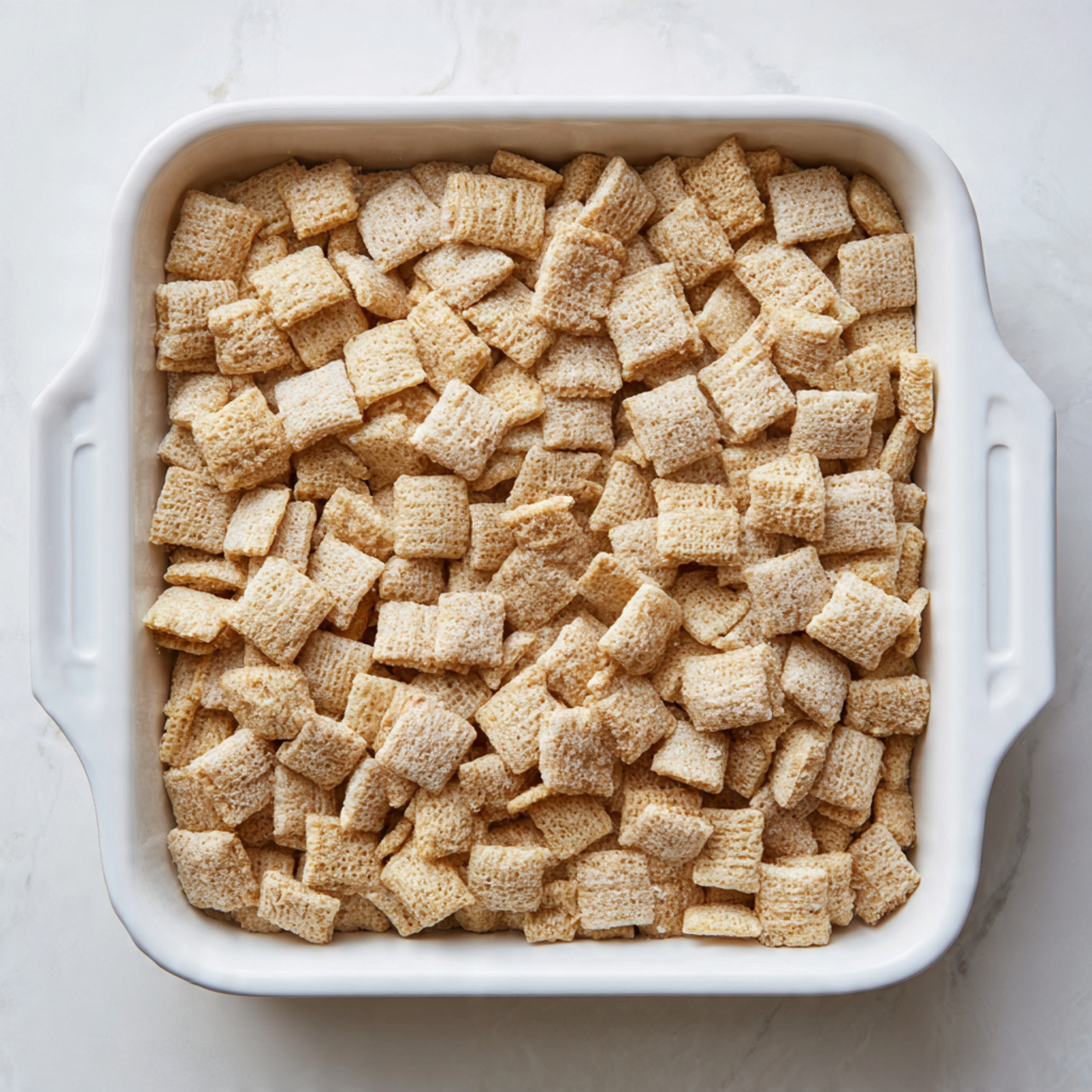 The image shows a large rectangular white baking dish filled with one layer of light golden square cereal pieces that are puffed and crispy with a slightly rough texture. The cereal pieces cover the entire dish evenly, with no additional layers or toppings visible. The dish sits on a white marbled surface with a soft light coming from the side, highlighting the pale beige colors of the cereal. Photo taken with an iphone --ar 4:5 --v 7