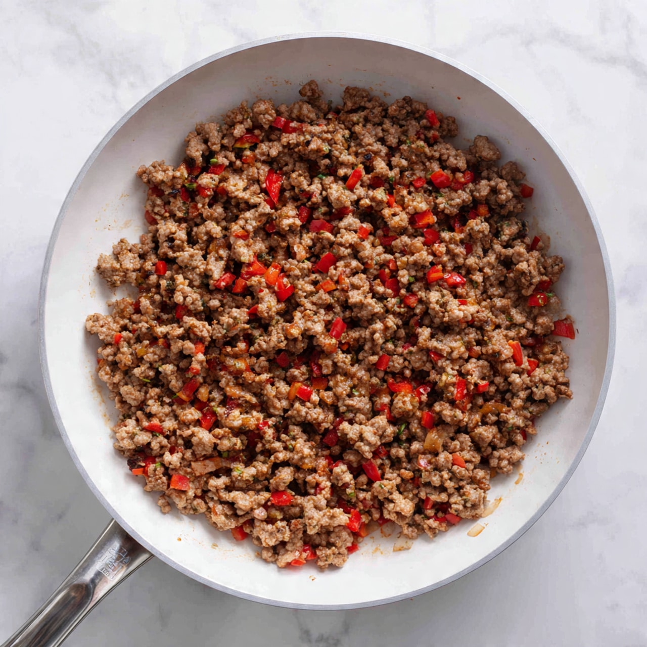The image shows a top-down view of a white frying pan filled with cooked ground meat mixed with small diced red bell peppers and bits of onion. The meat is a light brown color with a crumbly texture, evenly spread inside the pan. The red bell peppers add bright spots of color throughout, and the onions can be seen as small translucent pieces mixed in. The handle of the pan is silver and slightly curves upwards. The pan rests on a surface with a white marbled texture. photo taken with an iphone --ar 4:5 --v 7
