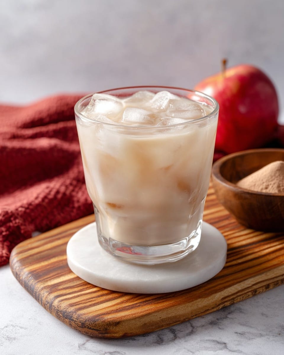 A clear glass filled with a pale creamy drink with many ice cubes inside, showing layers of white and light beige around the ice. The glass sits on a round white marble coaster, placed on a wooden cutting board with smooth grain patterns. In the background, there is a small wooden bowl with light brown powder and a red apple, along with a folded red cloth on a white marbled surface. photo taken with an iphone --ar 4:5 --v 7