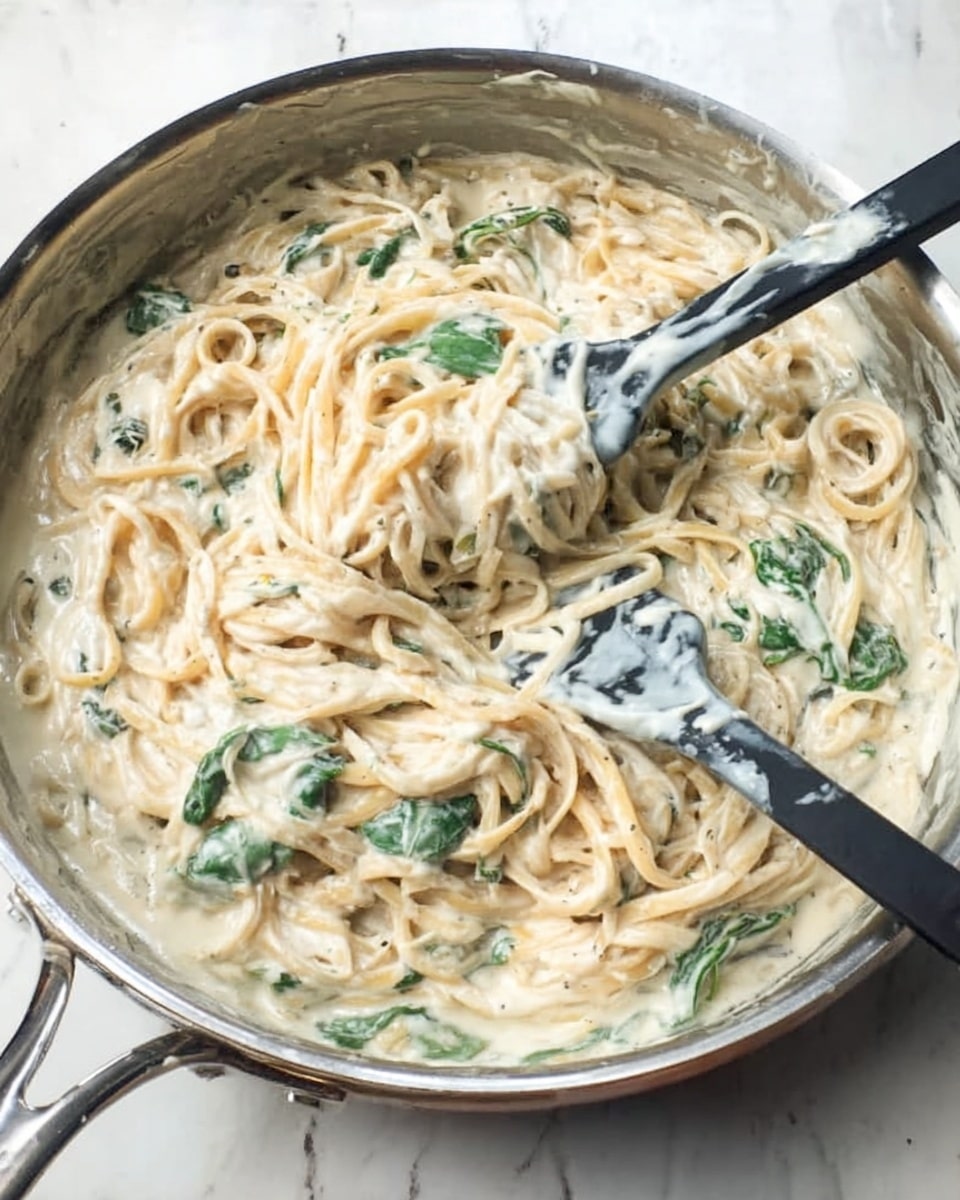 A silver pan filled with creamy pasta noodles coated in a thick white sauce mixed with pieces of green leafy vegetables. Two black and white spatulas rest inside the pan, one slightly lifted showing more sauce and noodles stuck to it. The pan is placed on a white marbled surface. The sauce looks smooth and rich with a slight shine. photo taken with an iphone --ar 4:5 --v 7