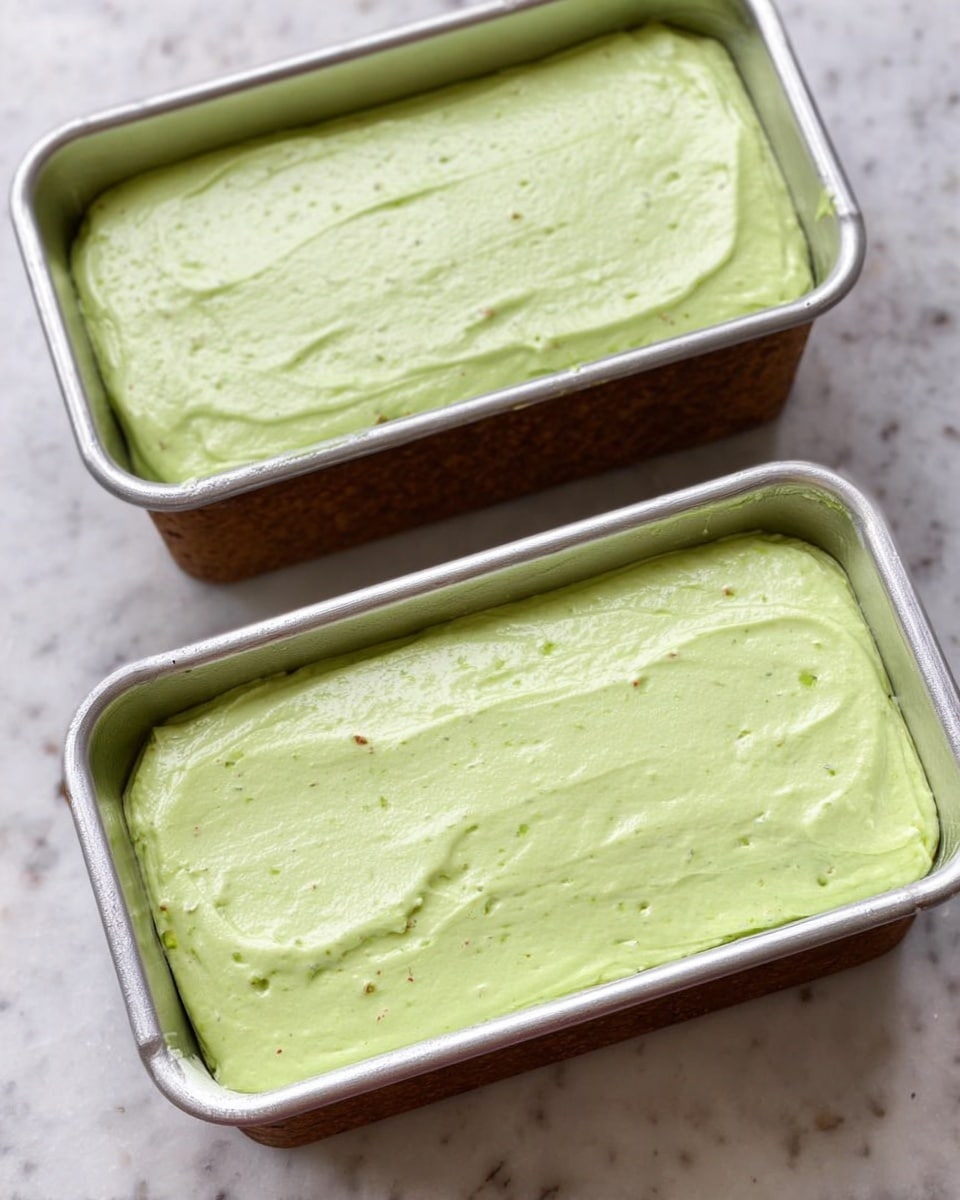 Two silver baking pans filled with smooth light green batter that looks creamy and evenly spread across the surface. Each pan is placed on a white marbled table, showing the batter's soft texture with some small bits inside. The pans are rectangular with rounded edges and the batter fills most of the space inside. Photo taken with an iphone --ar 4:5 --v 7