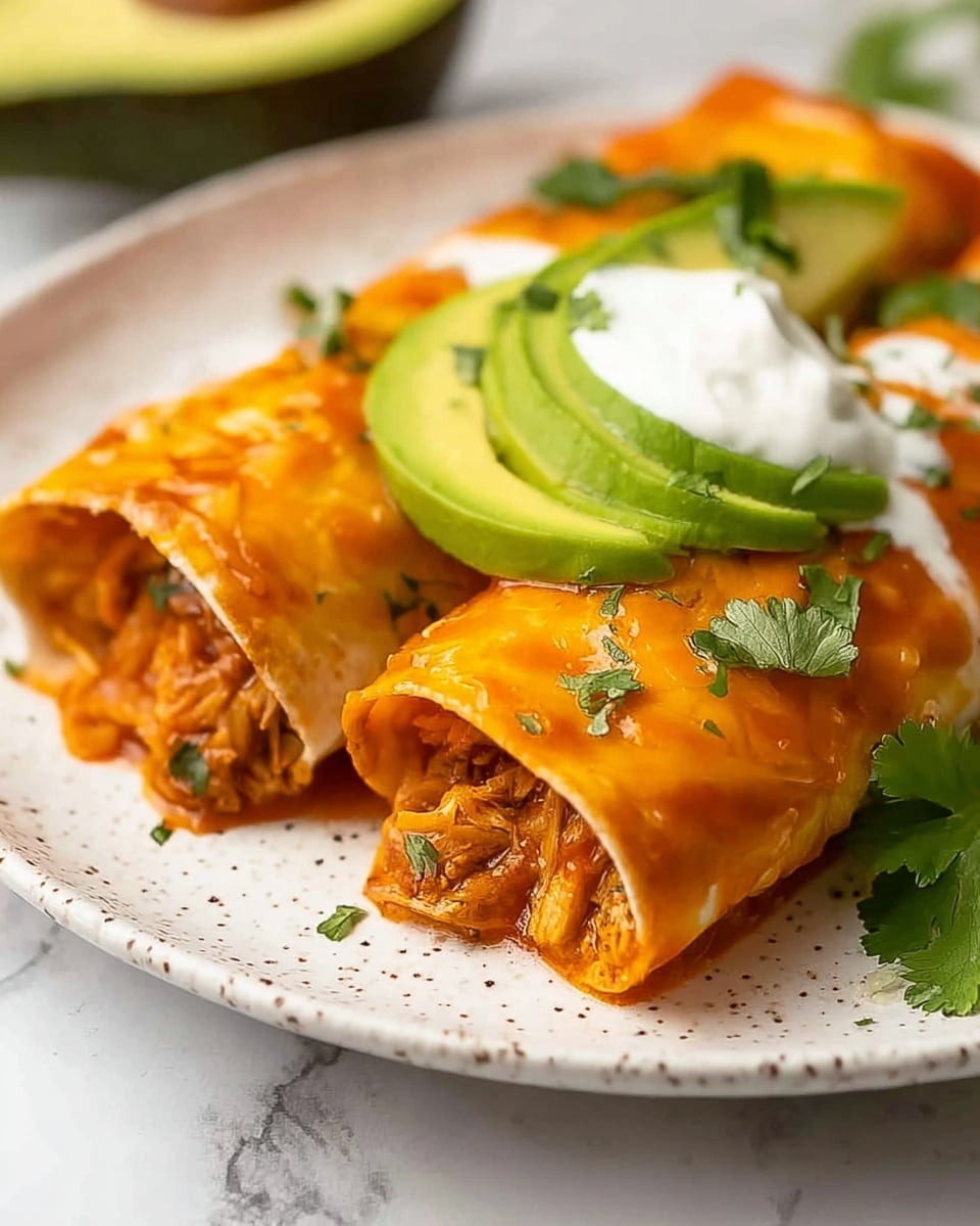Two rolled orange tortillas covered with a shiny sauce are placed side by side on a white speckled plate. Inside the tortillas, there are visible layers of light brown shredded filling mixed with bits of green herbs. On top of the rolls, there are two slices of light green avocado stacked, a dollop of white sour cream, and green cilantro leaves for garnish. The background shows a white marbled surface with a blurred avocado half in the foreground. photo taken with an iphone --ar 4:5 --v 7