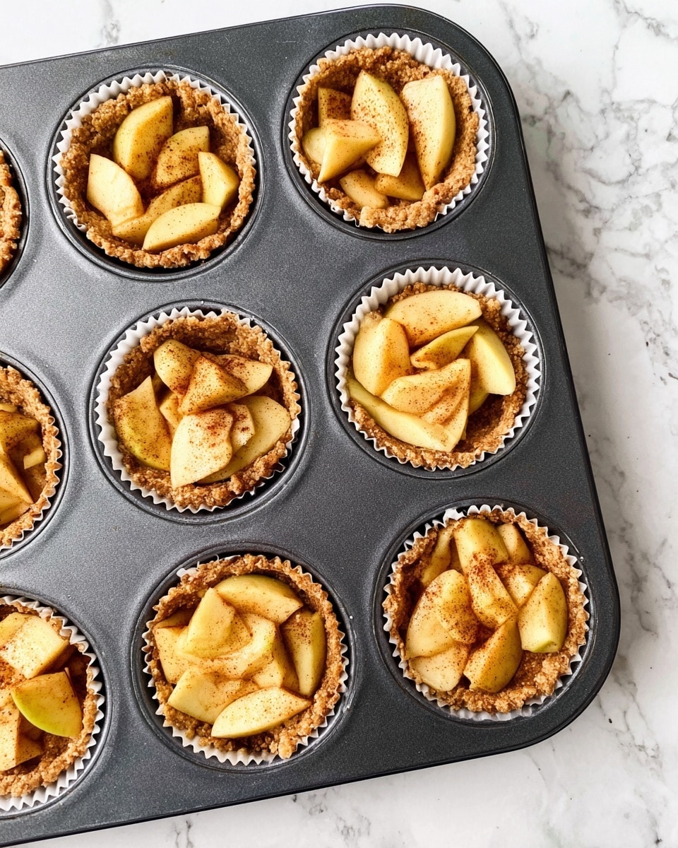 The image shows six mini apple tarts placed in a dark gray metal muffin tray on a white marbled surface. Each tart has two layers: a bottom layer of light brown crumbly crust inside white paper liners, and a top layer of thinly sliced apple pieces with a light yellow color sprinkled with brown cinnamon powder. The apple slices are arranged loosely and overlap in the center of each tart, showing a soft, slightly shiny texture. Photo taken with an iphone --ar 4:5 --v 7