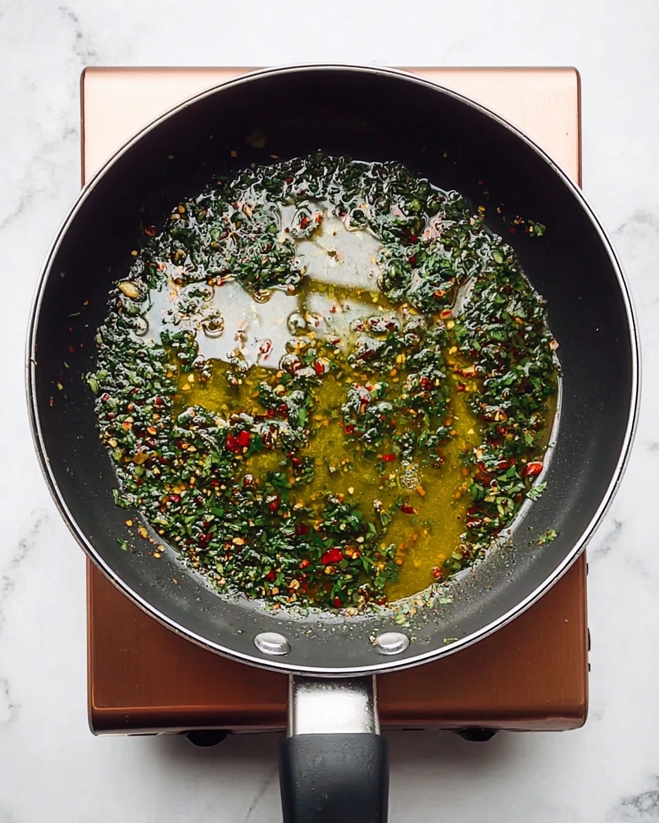 A large dark gray frying pan filled with a big nest of pale yellow spaghetti noodles, mixed evenly with small green parsley flakes and tiny red chili pieces, creating a colorful contrast throughout the pasta. Two wooden spoons rest inside the pan, one of them lifting a twisted bunch of noodles to show their smooth and glossy texture. The pan is set on a white marbled surface with a hint of the stove visible below. photo taken with an iphone --ar 4:5 --v 7
