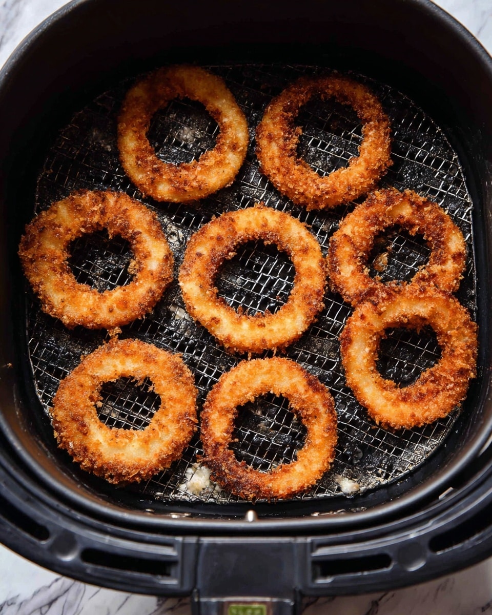 Seven golden brown onion rings with a crispy texture are arranged inside a black air fryer basket. The basket has a mesh bottom that shows some oil spots and crumbs. The onion rings vary slightly in size, with a crunchy outer layer that shows small uneven bits of breading. The light inside the basket highlights the warm fried tones of the rings. The background outside the basket is a white marbled texture. Photo taken with an iphone --ar 4:5 --v 7