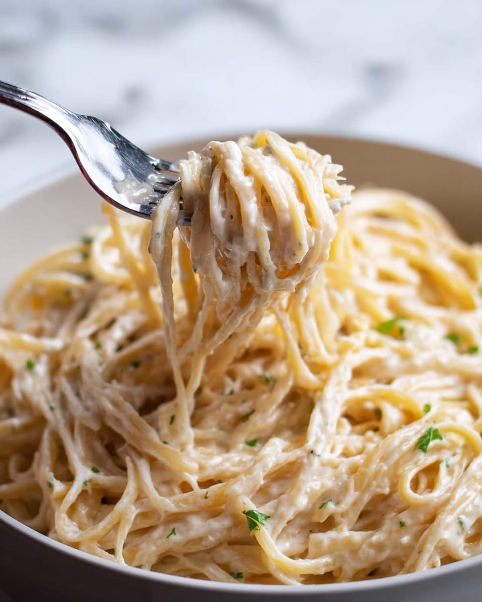 A close-up view shows creamy pasta coated in a light white sauce with small green herb bits, piled loosely in a white bowl. A silver fork lifts some strands of the pale golden noodles with a smooth, rich texture, twisting them near the center of the image. The pasta looks soft and well mixed with the sauce, giving a slightly glossy appearance. The background features a white marbled surface, creating a clean, bright setting for the dish. photo taken with an iphone --ar 4:5 --v 7