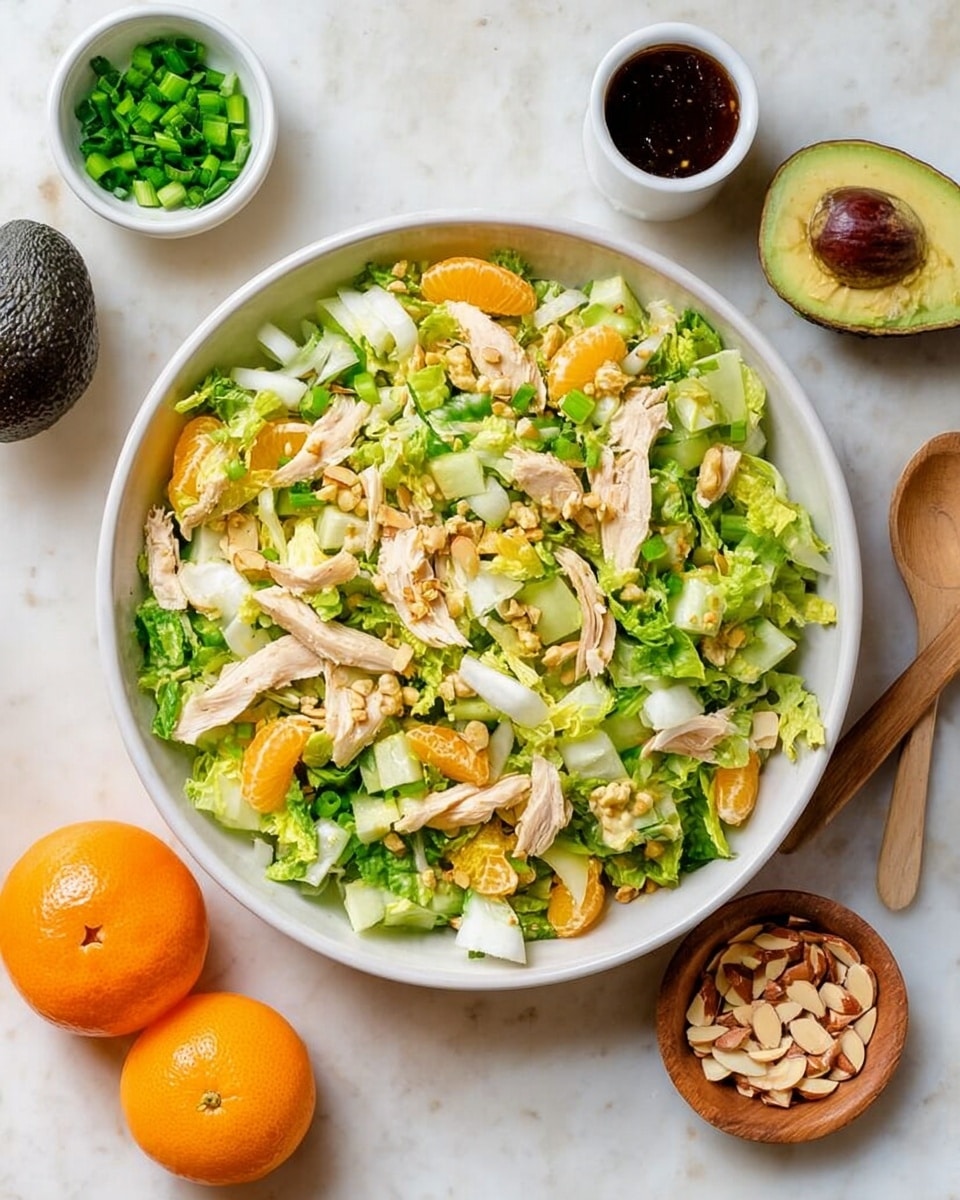 A large white bowl filled with a fresh salad made of chopped light green and yellow lettuce, white slices of celery, small dark green cucumber pieces, and light brown shredded chicken pieces mixed throughout. Around the bowl, there is a small white bowl of bright orange mandarin slices at the bottom left, a small white bowl with chopped green onions at the top center, a small round wooden dish with light brown sliced almonds at the bottom right, and a small white bowl with dark brown salad dressing and a wooden spoon near the top right. Half an avocado with a brown seed inside lies to the left side on a white marbled surface. Photo taken with an iphone --ar 4:5 --v 7