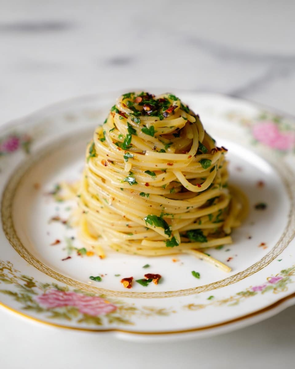 A small, neat stack of spaghetti sits in the center of a white plate with delicate floral and gold trim details. The pasta is yellowish, glistening lightly and mixed with finely chopped green herbs and red chili flakes scattered on top and around the base. The noodles are twirled to form a round, elevated mound, and the background features a clean white marbled texture. Photo taken with an iphone --ar 4:5 --v 7
