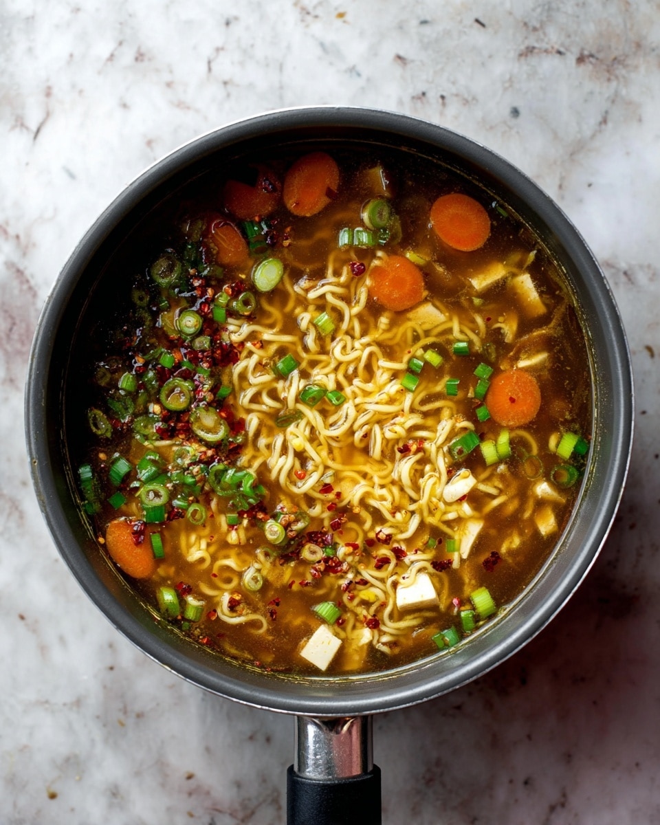 A grey pot filled with hot soup sits on a white marbled surface. The soup has several layers: a clear brown broth with floating thin wavy yellow noodles, small round orange carrot slices, small white rectangular pieces of tofu, chopped green onion slices, and bits of red chili flakes scattered on top. The pot has a black handle and is viewed from above. photo taken with an iphone --ar 4:5 --v 7