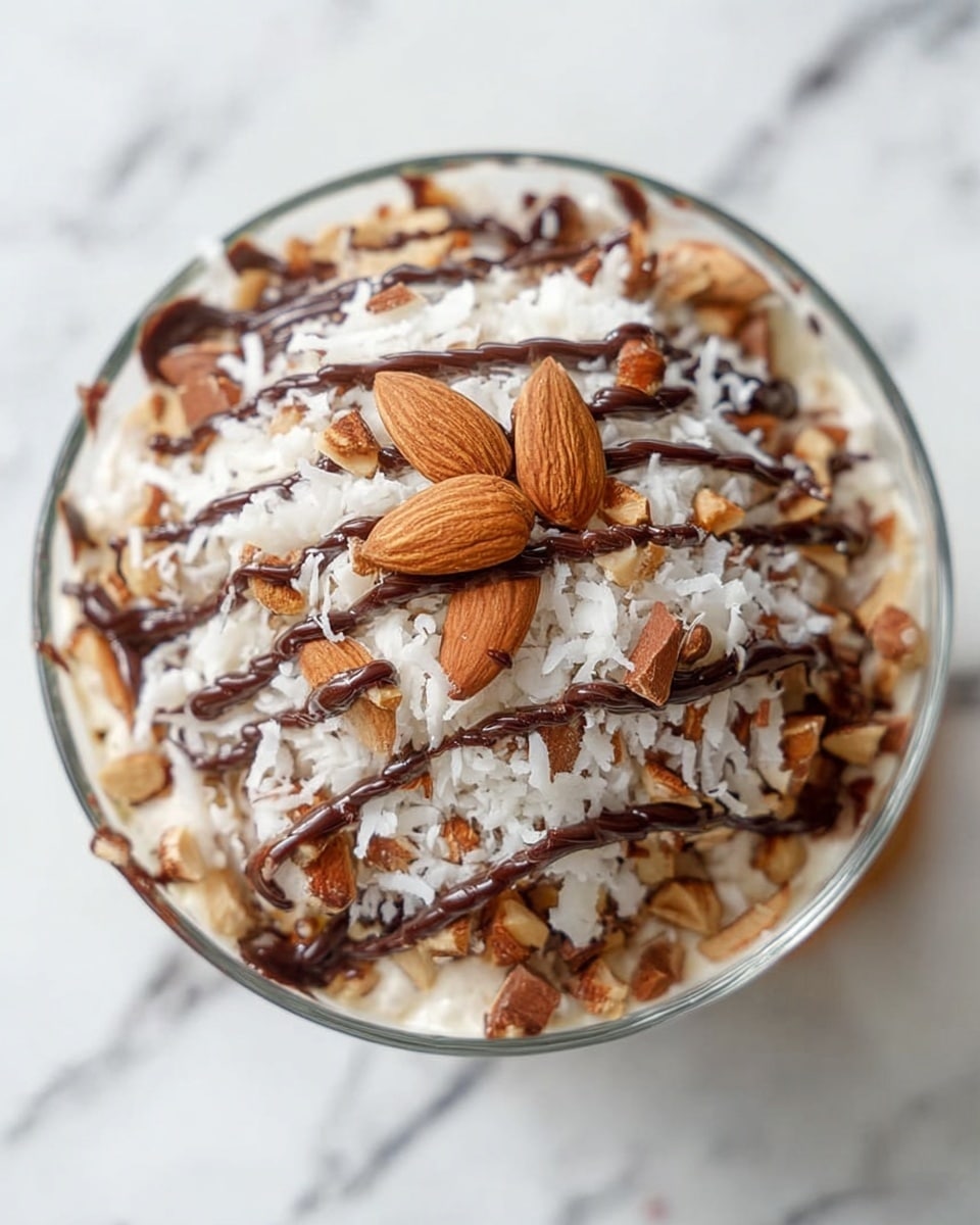 A dessert in a clear glass bowl filled with a creamy white base, topped with shredded white coconut spread evenly. On top of the coconut, there are several whole and pieces of brown almonds scattered all over. Thin, dark brown chocolate syrup is drizzled in lines across the top, adding contrast. A single whole almond is placed at the center on top, giving focus to the dish. The bowl is placed on a white marbled surface. photo taken with an iphone --ar 4:5 --v 7