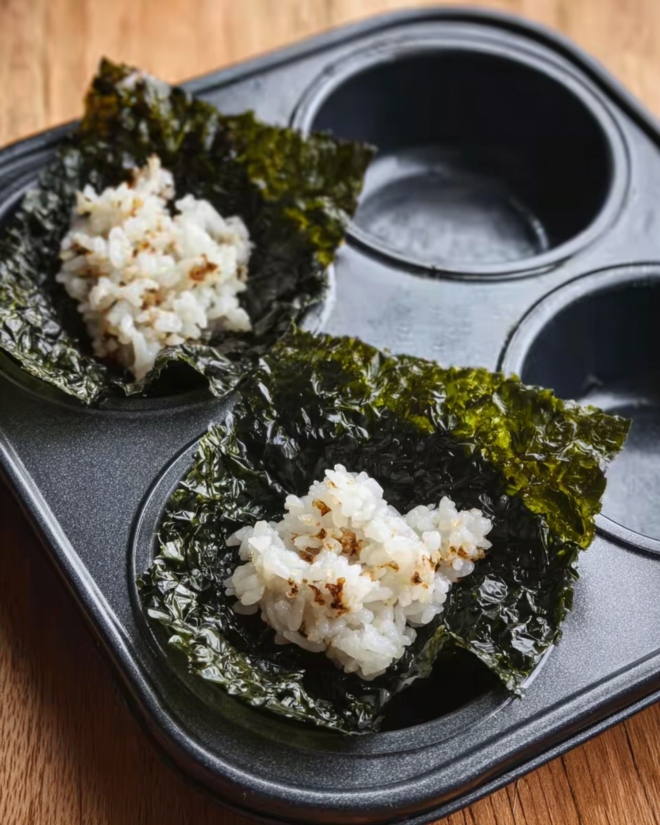 A black metal muffin tray holds two pieces of seaweed with rice on top, placed inside two cups of the tray; the seaweed is dark green with a crinkled texture, square-shaped and larger than the rice portions. The rice is white with some darker bits mixed in, slightly sticky and piled loosely in small clumps on the seaweed. The tray rests on a wooden surface. photo taken with an iphone --ar 4:5 --v 7