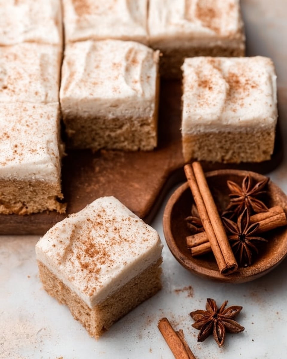 The image shows a close-up of square pieces of cake with two layers. The bottom layer is a light brown, soft cake with a slightly crumbly texture, and the top layer is thick, creamy white frosting sprinkled lightly with brown spice powder. One piece is slightly pulled away, showing the smooth frosting and fluffy cake inside. Next to the cake is a small wooden bowl with cinnamon sticks and star anise on a white marbled surface. Photo taken with an iphone --ar 4:5 --v 7