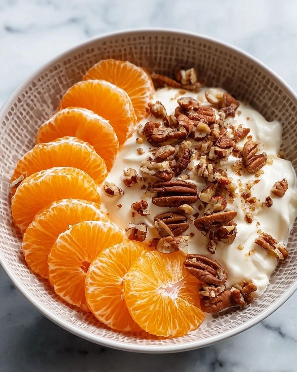 This dish is served in a white bowl with a dotted pattern on the outside, placed on a white marbled surface. Inside the bowl, there are two main layers: on one side, bright orange mandarin segments are carefully arranged in a semi-circle, showing their smooth, shiny texture. The other side has a generous dollop of white creamy mixture topped with a scattering of brown pecans and smaller crushed nut pieces, adding a rough texture contrast. The bright orange and white colors stand out clearly against the bowl and surface. Photo taken with an iphone --ar 4:5 --v 7