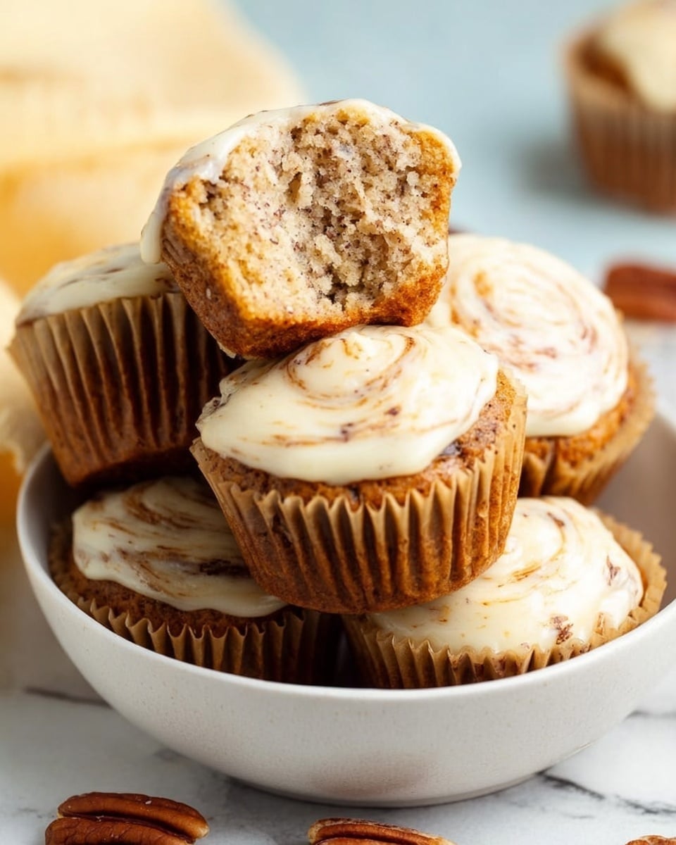 A close-up of a white bowl filled with six muffins topped with creamy white frosting that has light brown swirls. One muffin is held above the others, showing its inside texture which is light brown with small darker specks. The muffins have light brown paper wrappers and the bowl is placed on a white marbled surface with a few pecans scattered nearby. The lighting is soft and natural, highlighting the moist texture of the muffins and glossy frosting. Photo taken with an iphone --ar 4:5 --v 7