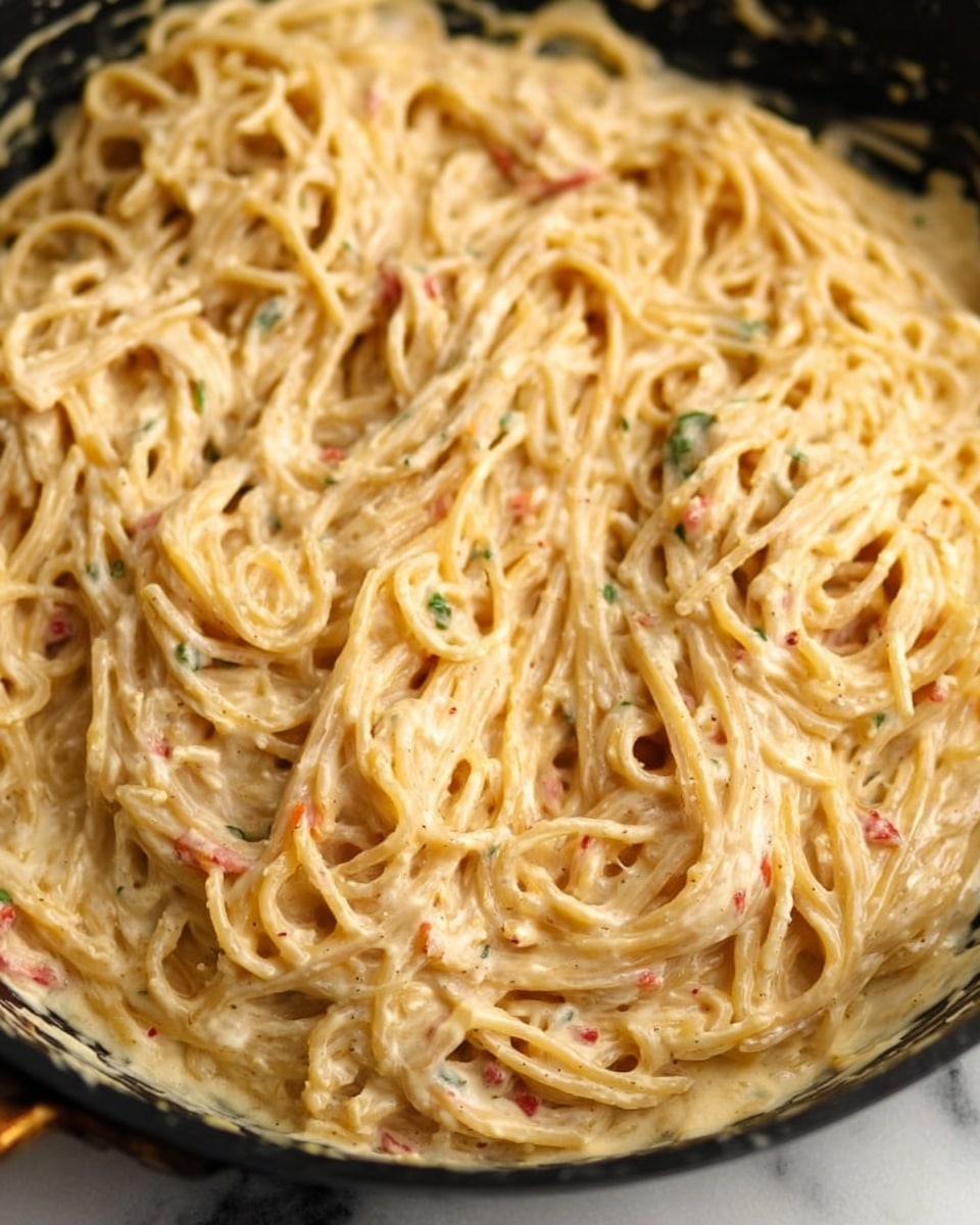 The image shows a close-up view of creamy pasta in a pan. The pasta strands are long and thin, coated in a thick, light beige sauce with specks of red and green herbs spread evenly throughout. The sauce looks rich and smooth, covering every strand well. The pasta fills the pan with slight texture from the sauce and bits of ingredients, creating a uniform, dense pile of noodles. The edge of the black pan is partly visible under the pasta around the sides. The background is a white marbled texture. photo taken with an iphone --ar 4:5 --v 7