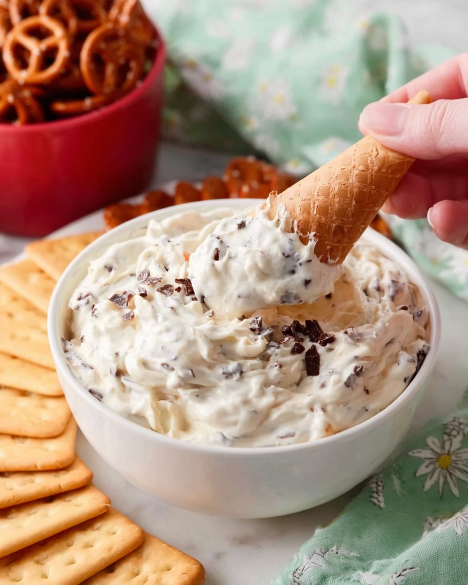 A white bowl filled with creamy white dip mixed with small dark bits, looking soft and thick. Around the bowl on a white marbled surface, there are rectangular light brown crackers standing upright and a red bowl full of pretzels in the background. A woman's hand is holding a small waffle cone, scooping up some of the dip from the bowl. The setting has a light green cloth with white floral patterns showing slightly behind the bowl. photo taken with an iphone --ar 4:5 --v 7
