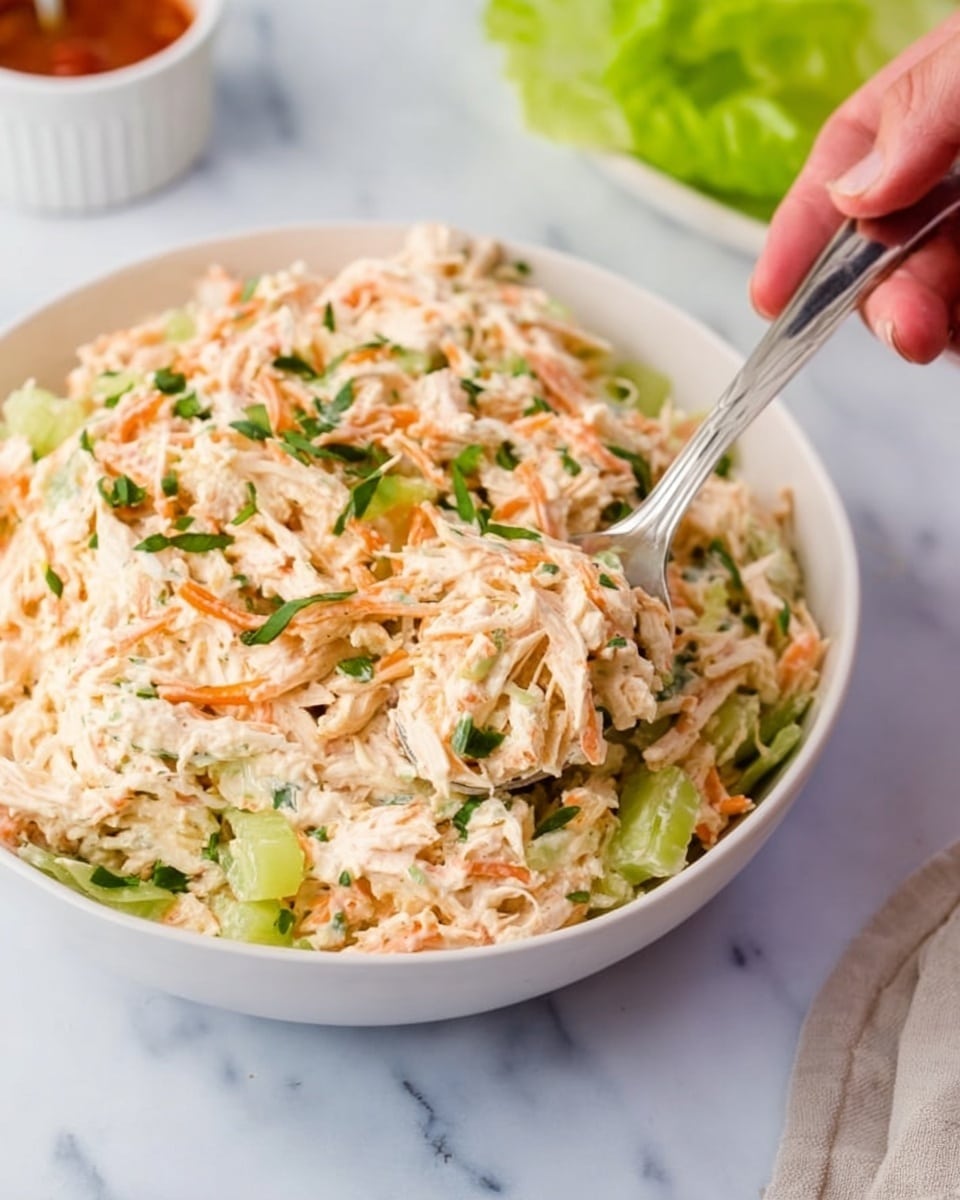 A white bowl filled with a creamy shredded chicken salad mixed with thinly sliced green celery and orange carrot shreds, with small bits of chopped green herbs sprinkled on top; the salad has a soft and smooth texture with visible fibrous pieces from the chicken; a woman's hand holding a silver spoon scooping some of the salad from the right side of the bowl; the surface below is white marble, and there is a hint of a green lettuce leaf and a white ramekin with a red sauce in the background. photo taken with an iphone --ar 4:5 --v 7